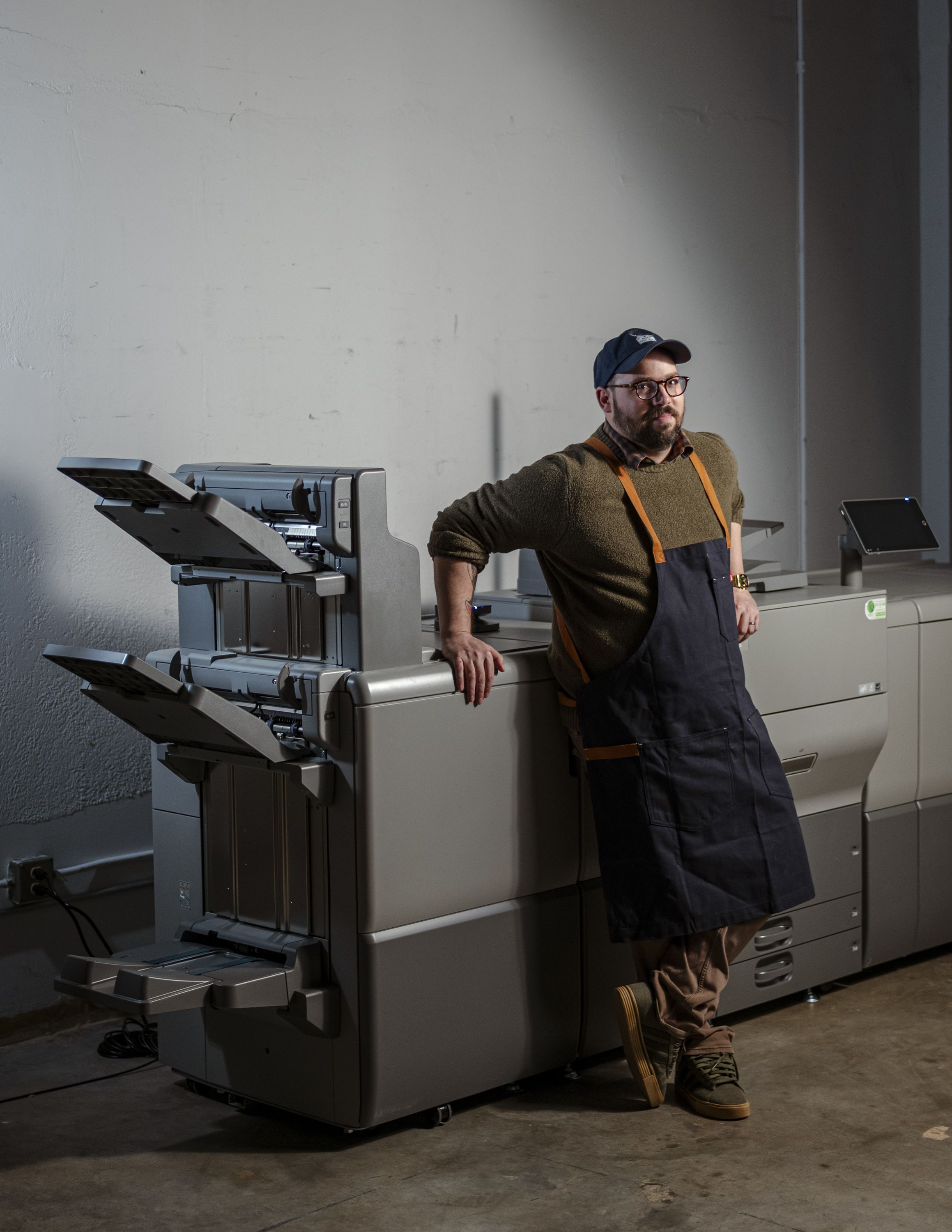 Man with glasses, cap, olive green sweater, and dark apron leans against large gray printing machine in industrial space with concrete floor and white wall.