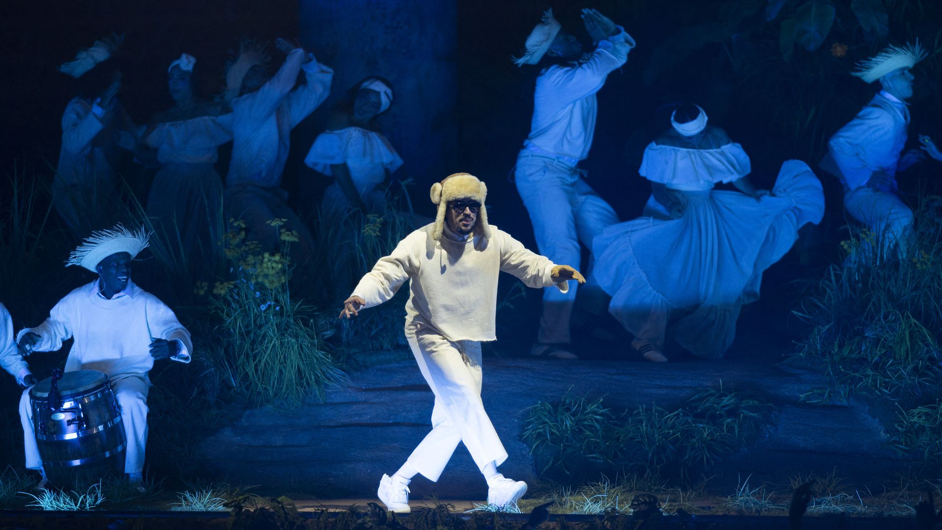 A man in a mostly white outfit and a bushy hat with sunglasses dances slightly on a blue-hued stage. 