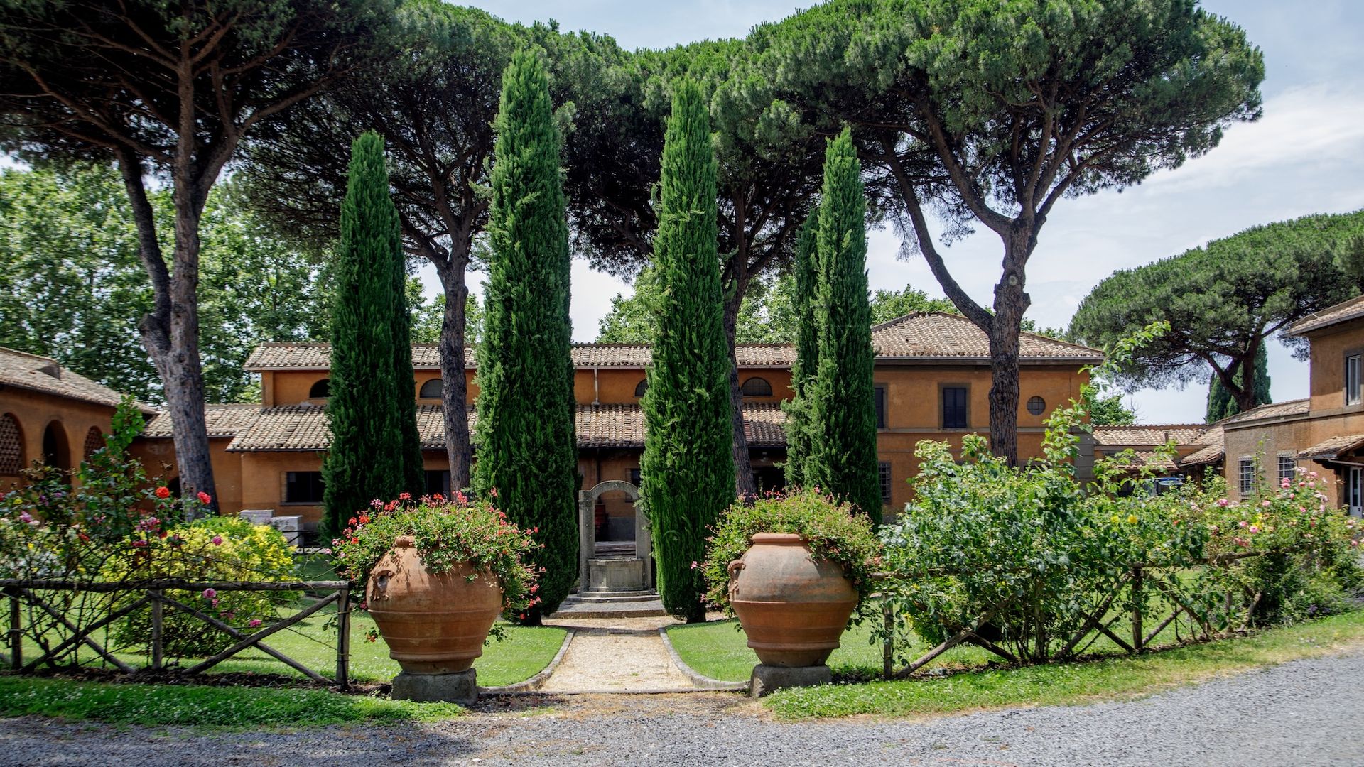 Mediterranean courtyard with tall cypress trees, large terracotta pots with flowering plants, surrounded by rustic orange buildings and umbrella pines under a clear sky.