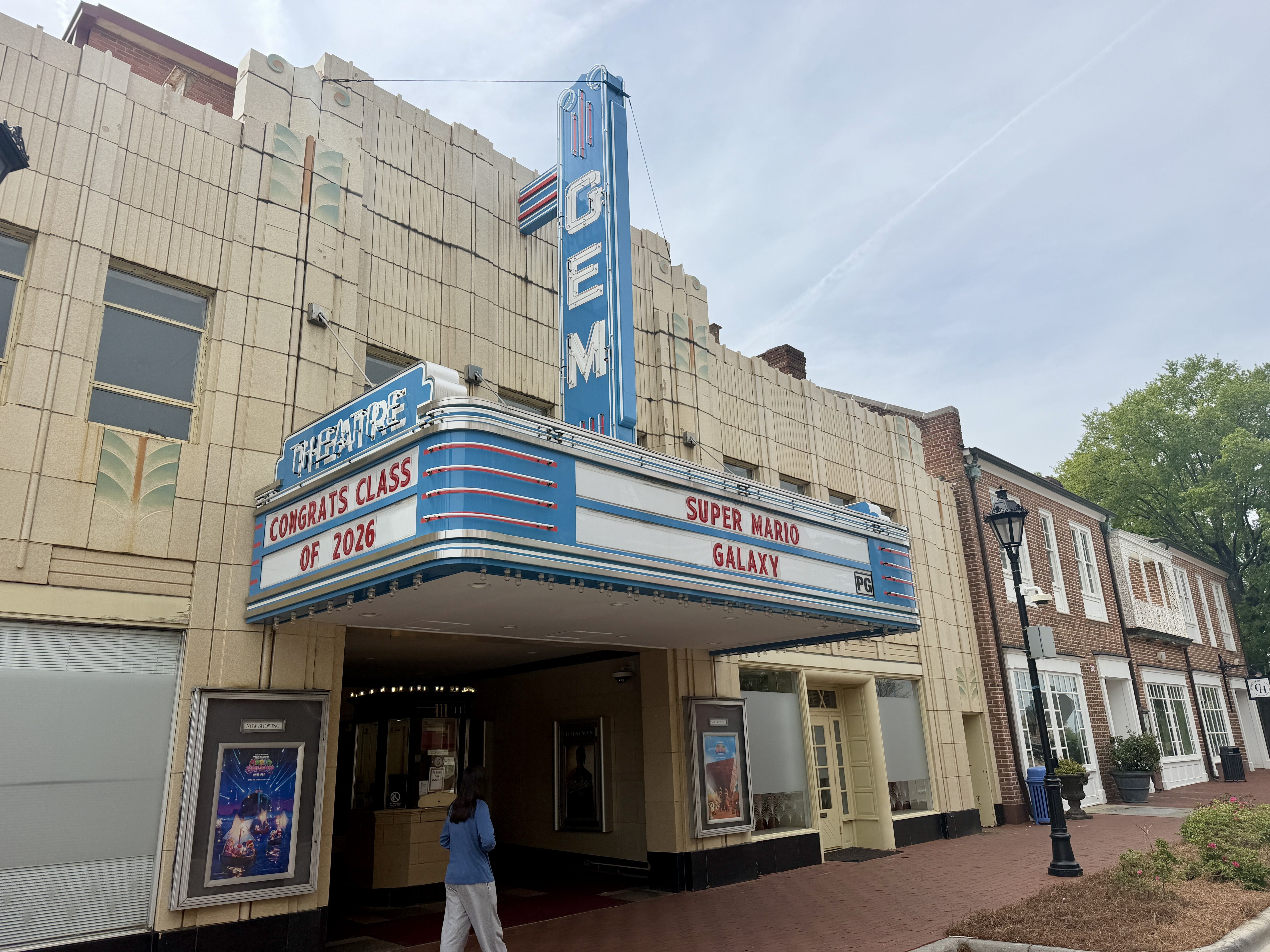 Retro theater marquee on a beige brick building with blue neon trim. It says CONGRATS CLASS OF 2026 and SUPER MARIO GALAXY. A person in a blue jacket walks along a brick sidewalk; trees in background.