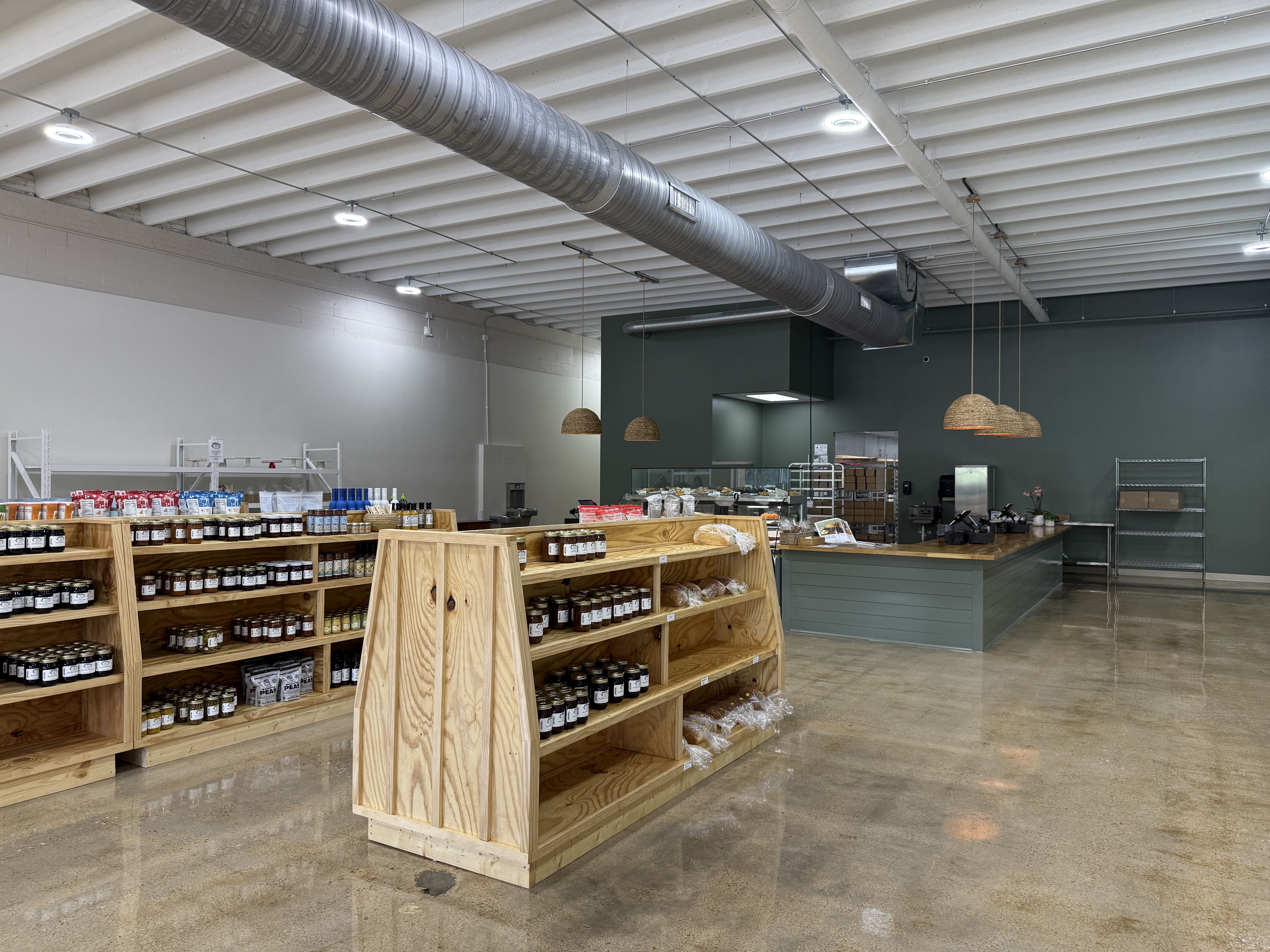 Interior of a modern store with wooden shelves stocked with jars, bread, and goods on a polished concrete floor. Green counter with wicker pendant lights hangs from a white ceiling with ductwork.