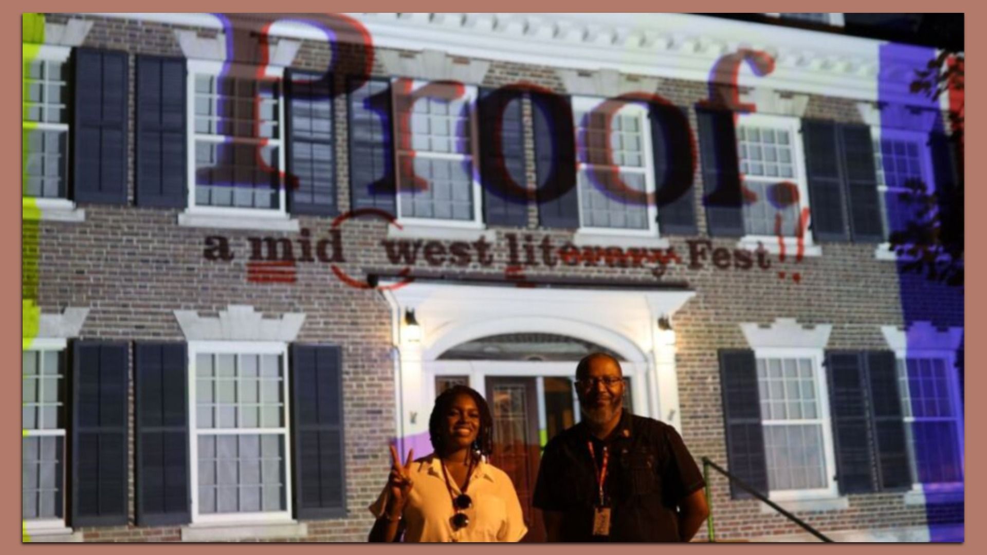 Two people stand smiling at night in front of a brick building with black shutters. A colorful projection shows the word "Proof" and "a mid west lit Fest!!" on the building.