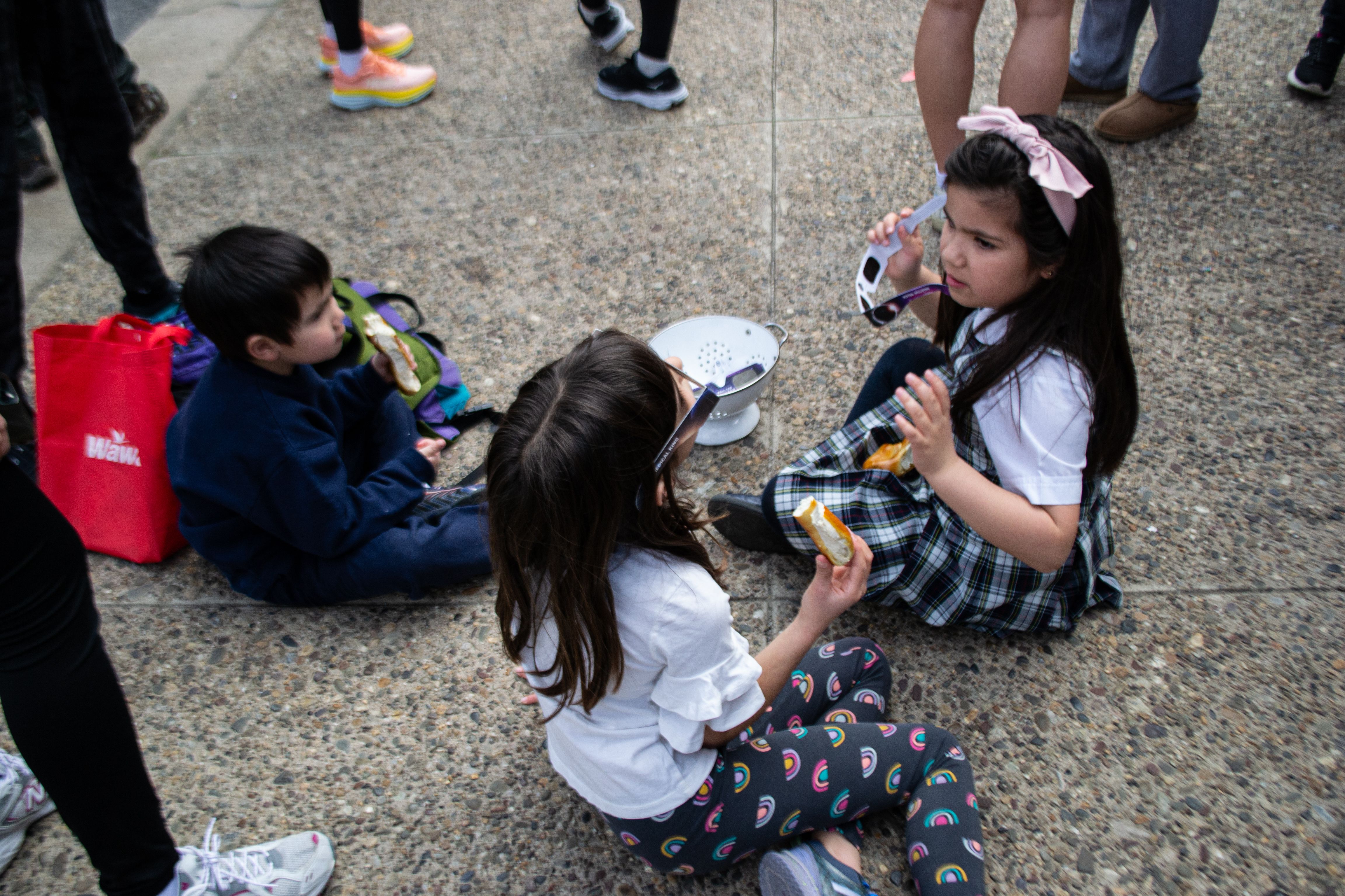 Children sit on the ground eating soft pretzels and holding eclipse glasses.