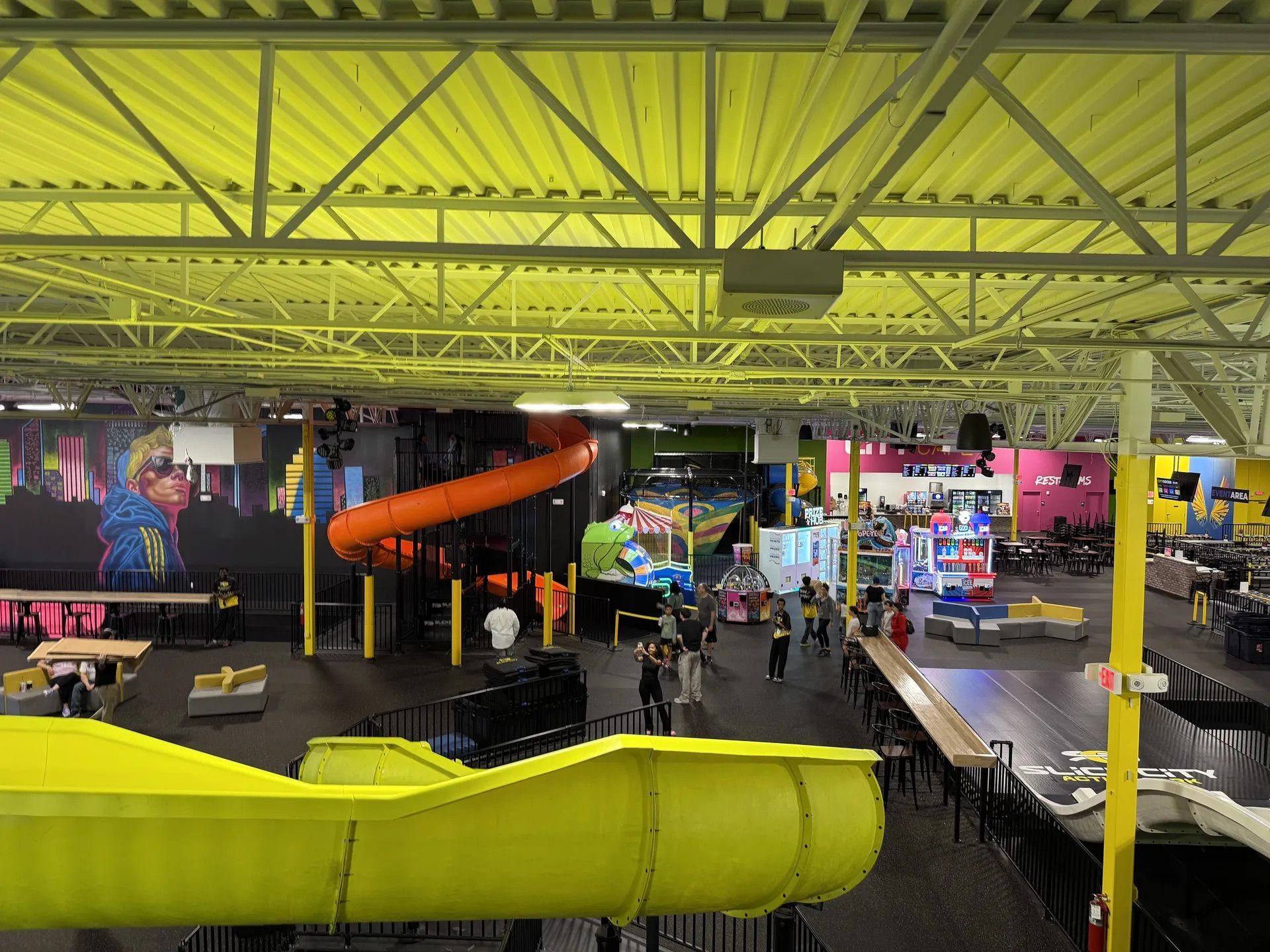 Slide Park. Slick City. Interior arcade and play area with a bright yellow ceiling and exposed truss. A large yellow slide curves in the foreground; an orange spiral slide, murals, arcade machines, and people in the background.