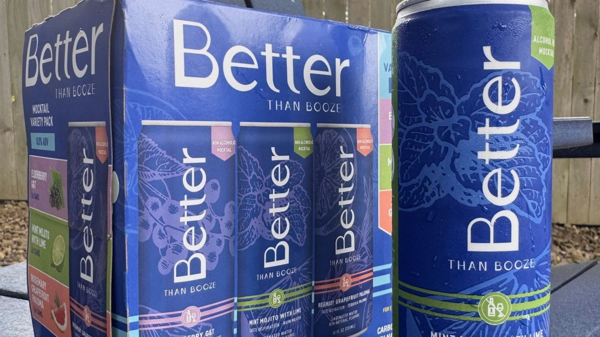 A six-pack of "Better Than Booze" non-alcoholic mocktails sits on a table with a can of Mint Mojito with Lime in front.