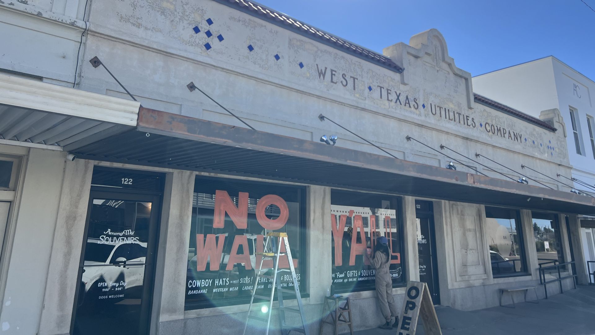 Sunny day over a weathered white storefront reading West Texas Utilities Company with blue diamond accents. A metal awning, large glass windows with pink lettering, and a person near the front door.
