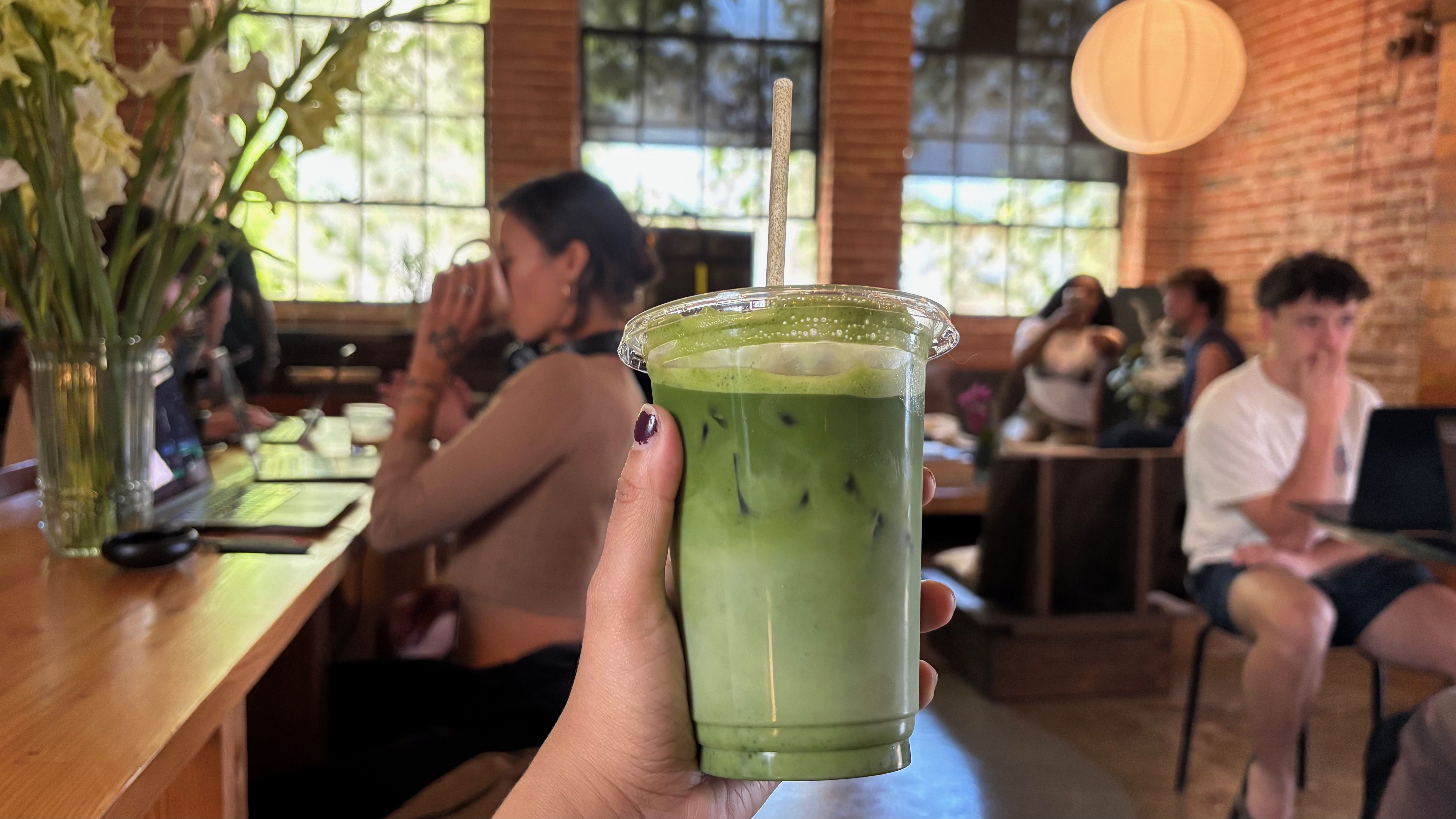 Hand with purple nails holds a clear cup of iced green matcha latte with a straw. Background shows a brick-walled cafe, large windows, a round lantern, flowers, and seated patrons.