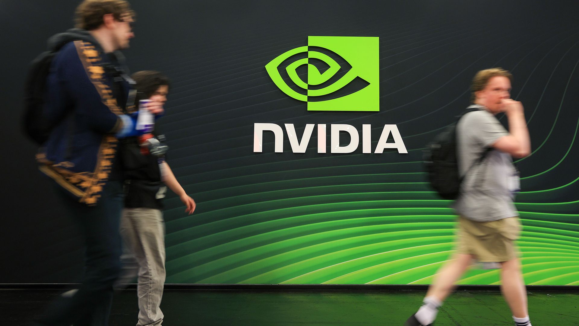 Blurred people walk past a dark wall featuring a bright green NVIDIA logo and the word NVIDIA in white, with curved green lines in the background.