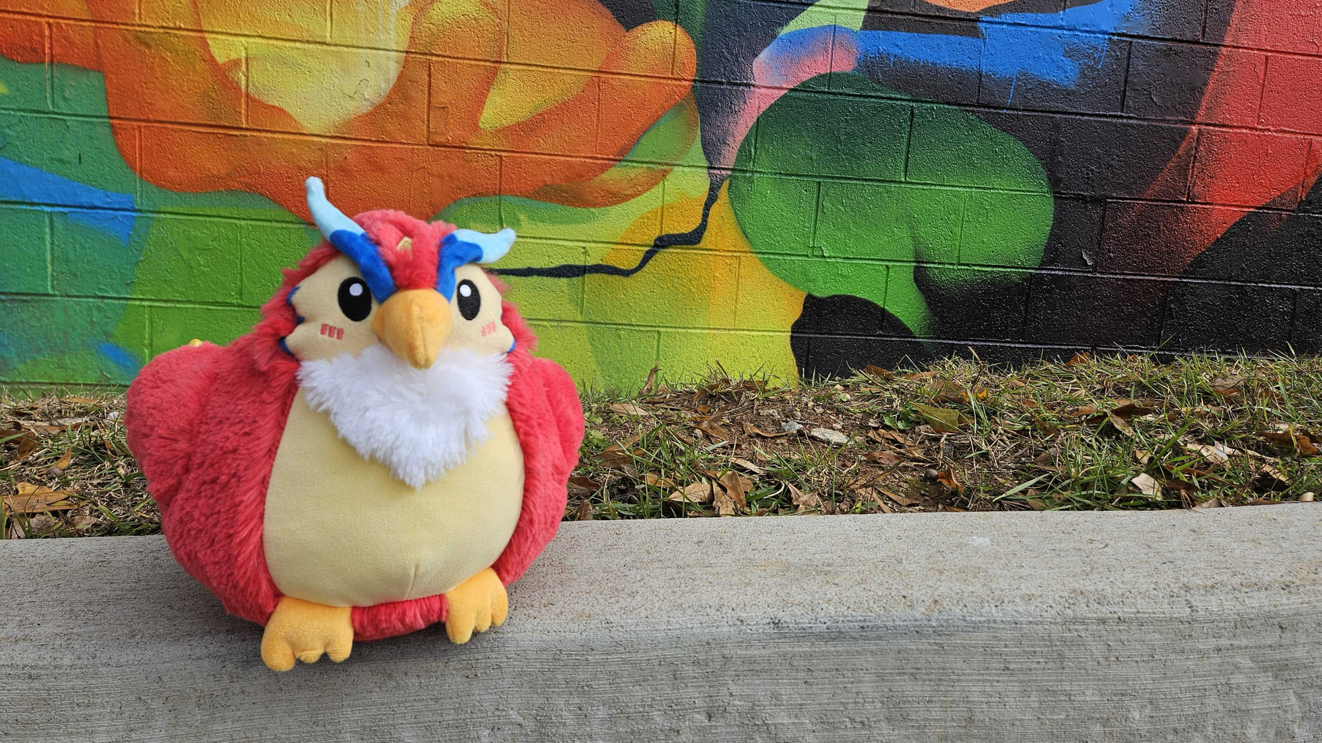 A stuffed toy Phoenix sits on a curb in front of a colorful mural