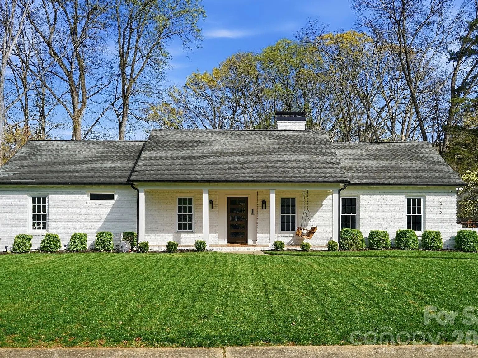 White brick ranch-style house with a dark gray roof, a centered front porch with two white columns, a wooden swing, neatly trimmed shrubs, and a lush green lawn under a blue sky.