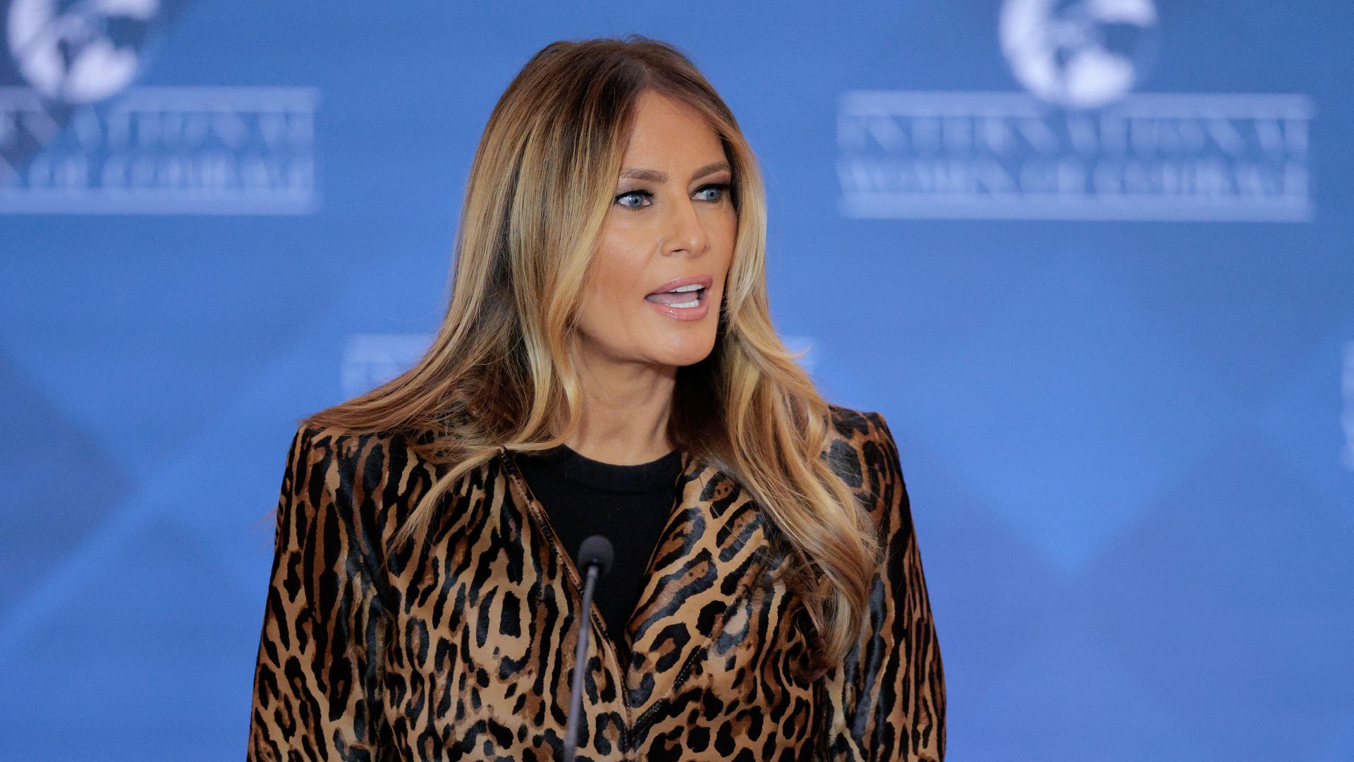 Melania Trump, wearing an animal print jacket, speaks into a microphone. A blue backdrop is behind her. 
