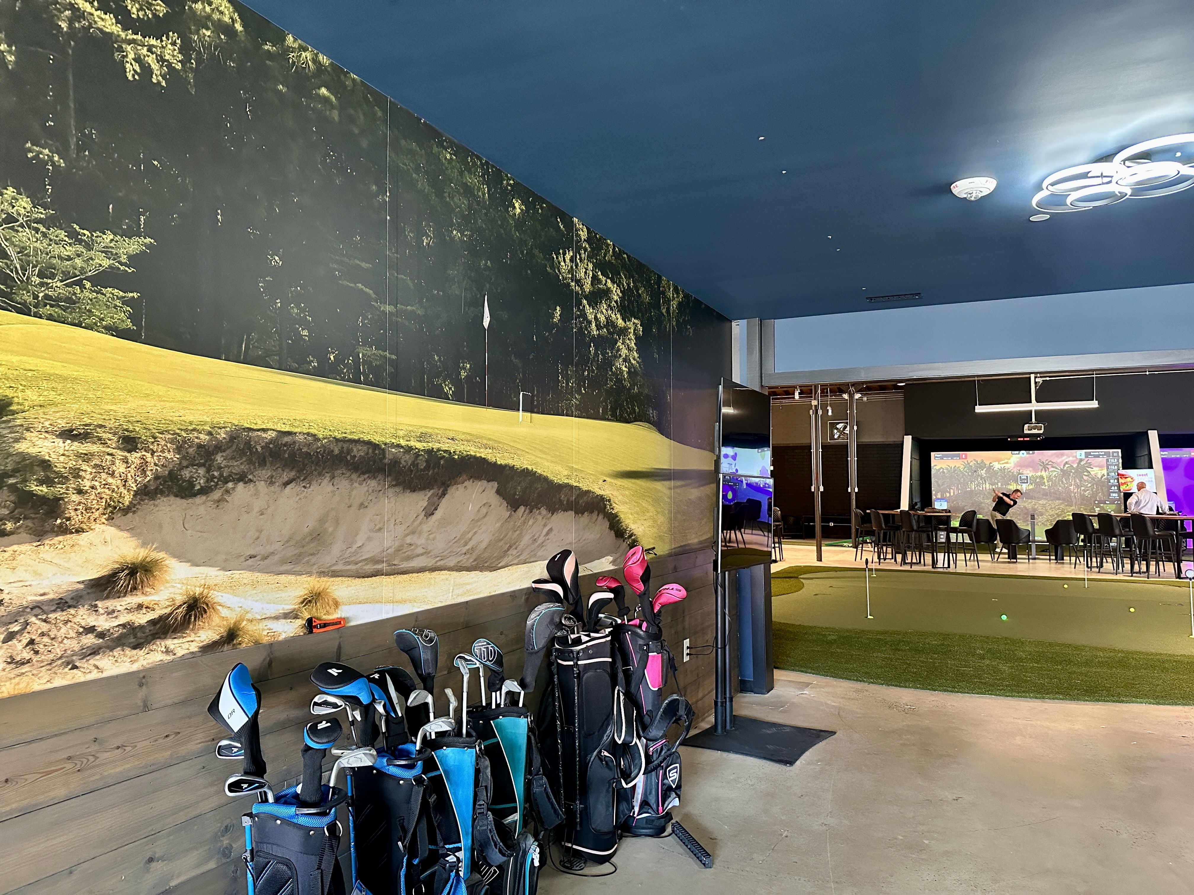 Indoor golf practice area with artificial turf, putting green, and seated players using golf simulators. Golf bags with clubs line the wall featuring a large golf course mural.