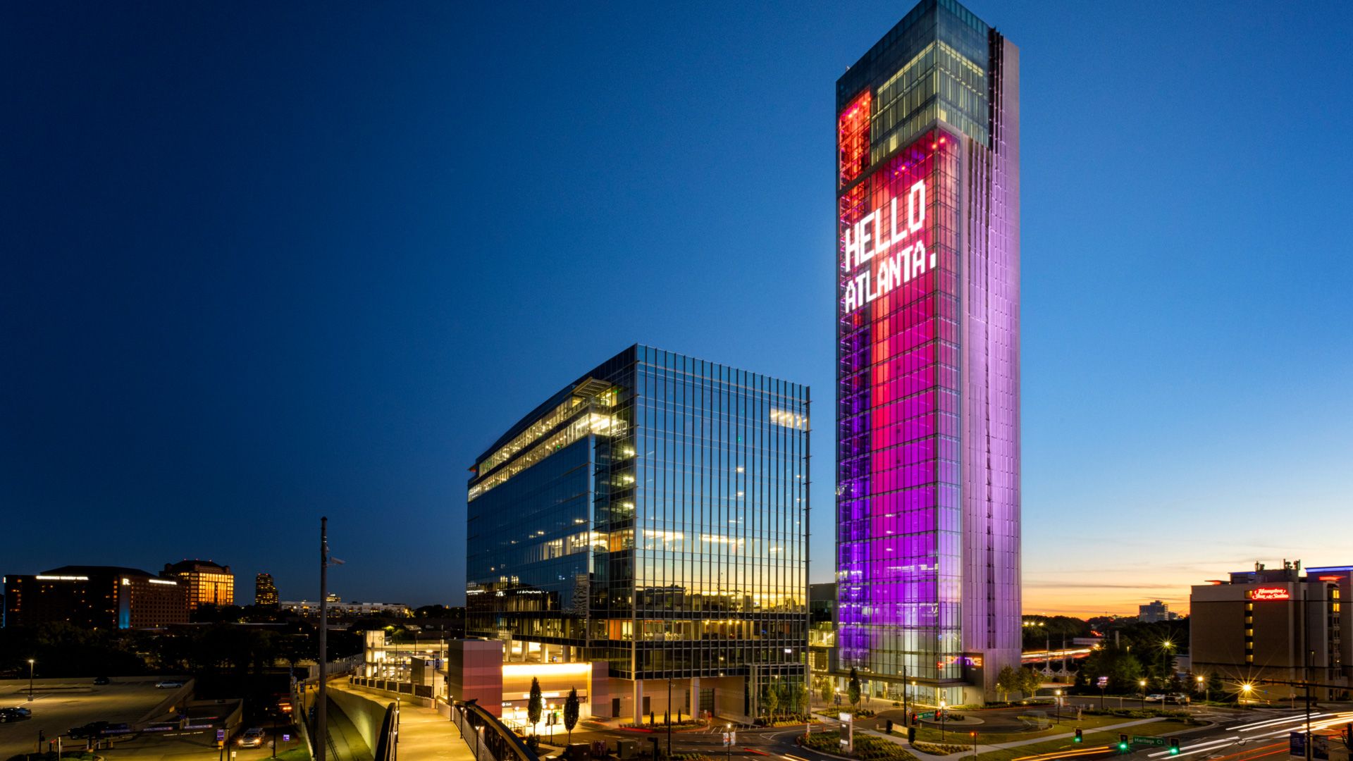 A tall tower illuminated in purple and red beams of light with a large digital sign saying "Hello, Atlanta"
