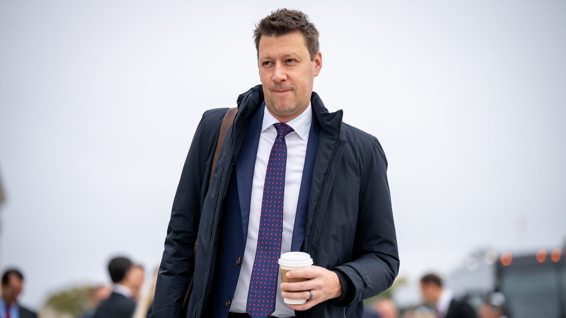 A man in a dark coat over a navy suit and red polka-dot tie walks outside, holding a takeaway coffee, with blurred people in the background.