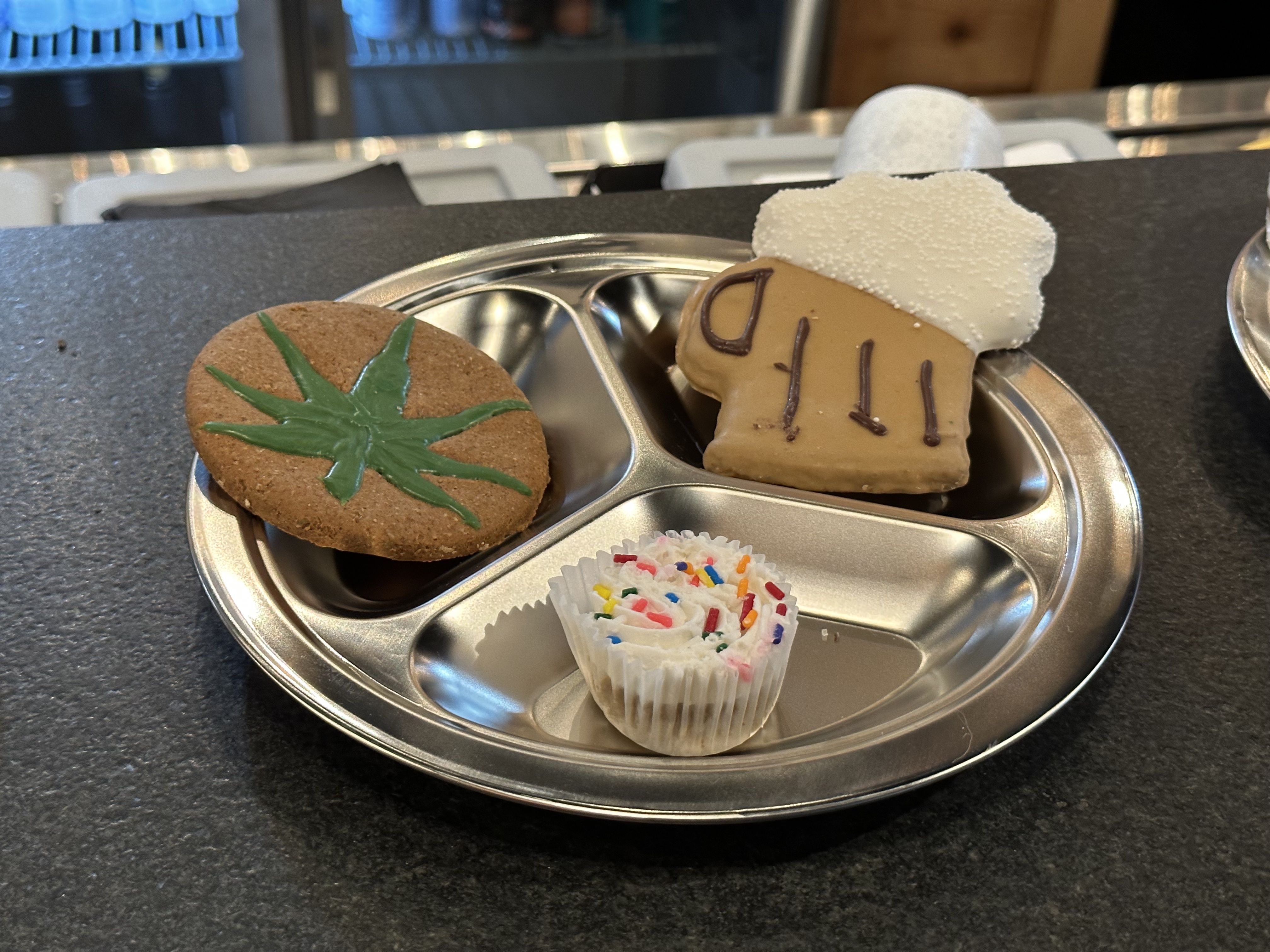 A metallic plate with three dog treats: One shaped and designed like an overflowing pint of beer, one with a green marijuana flower and one that's a white cupcake with rainbow sprinkles.