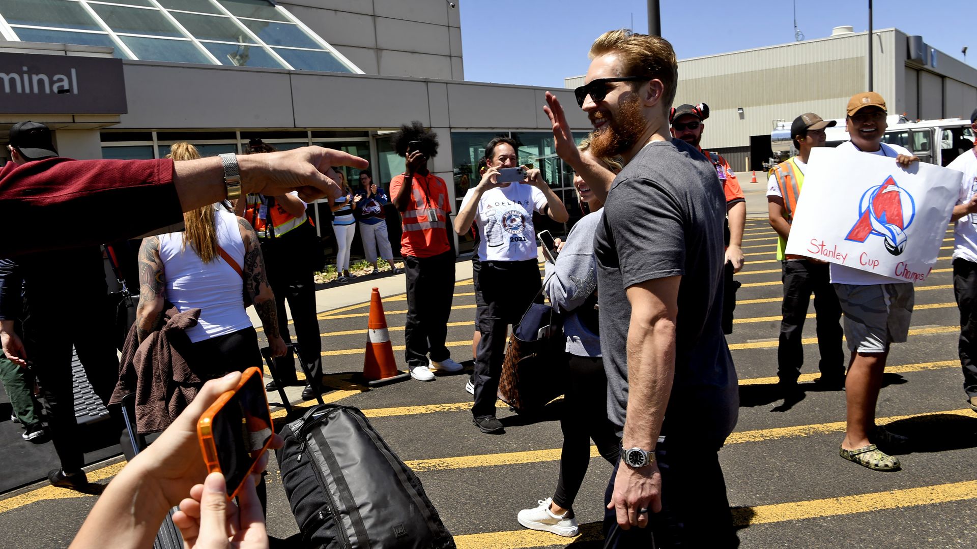 A man in a t shirt and sunglasses waves as he walks on an airport tarmac. 