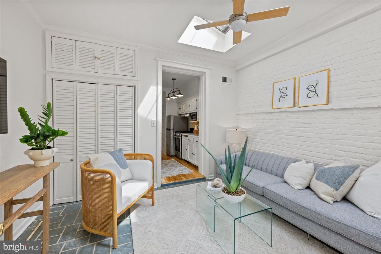 Bright living room with white brick wall, light gray sofa, wicker chair, glass coffee table with a potted plant, and a wooden ceiling fan; skylight above and doorway to a white kitchen ahead.
