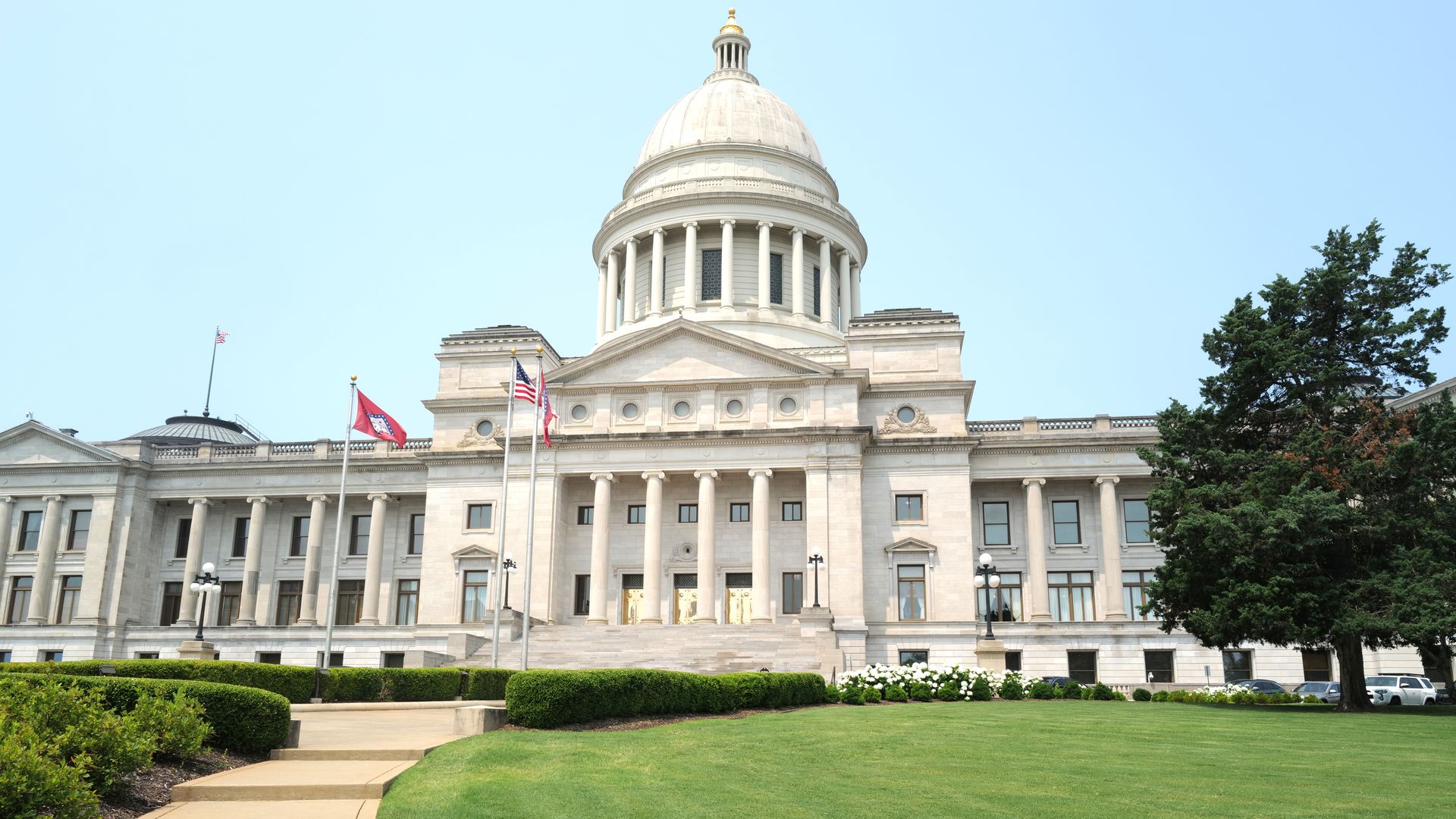 Wide view of a white neoclassical government building with a large domed roof, flanked by tall columns, flagpoles with American flags, and a green lawn under a clear blue sky.