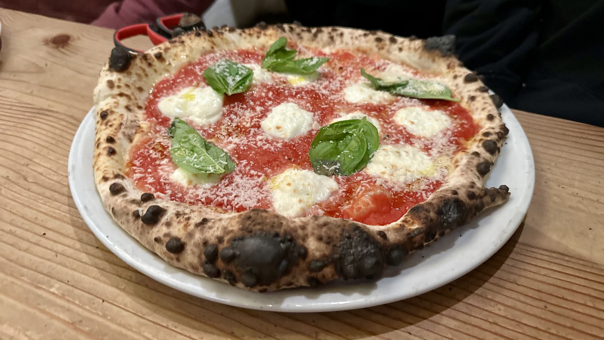 A pizza with puffed and blistered crust and dobs of buffalo mozzerella and whole basil leaves on a red sauce, sitting on a white plate on a wood table.