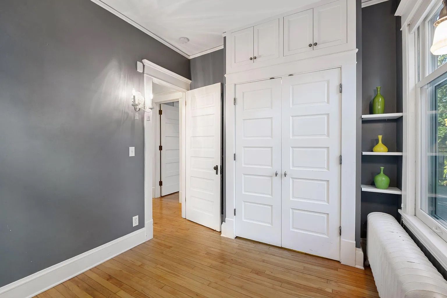 Bright room with gray walls, white paneled doors and shelves holding three green vases, hardwood floor, a window with white trim, and a white radiator below the window.