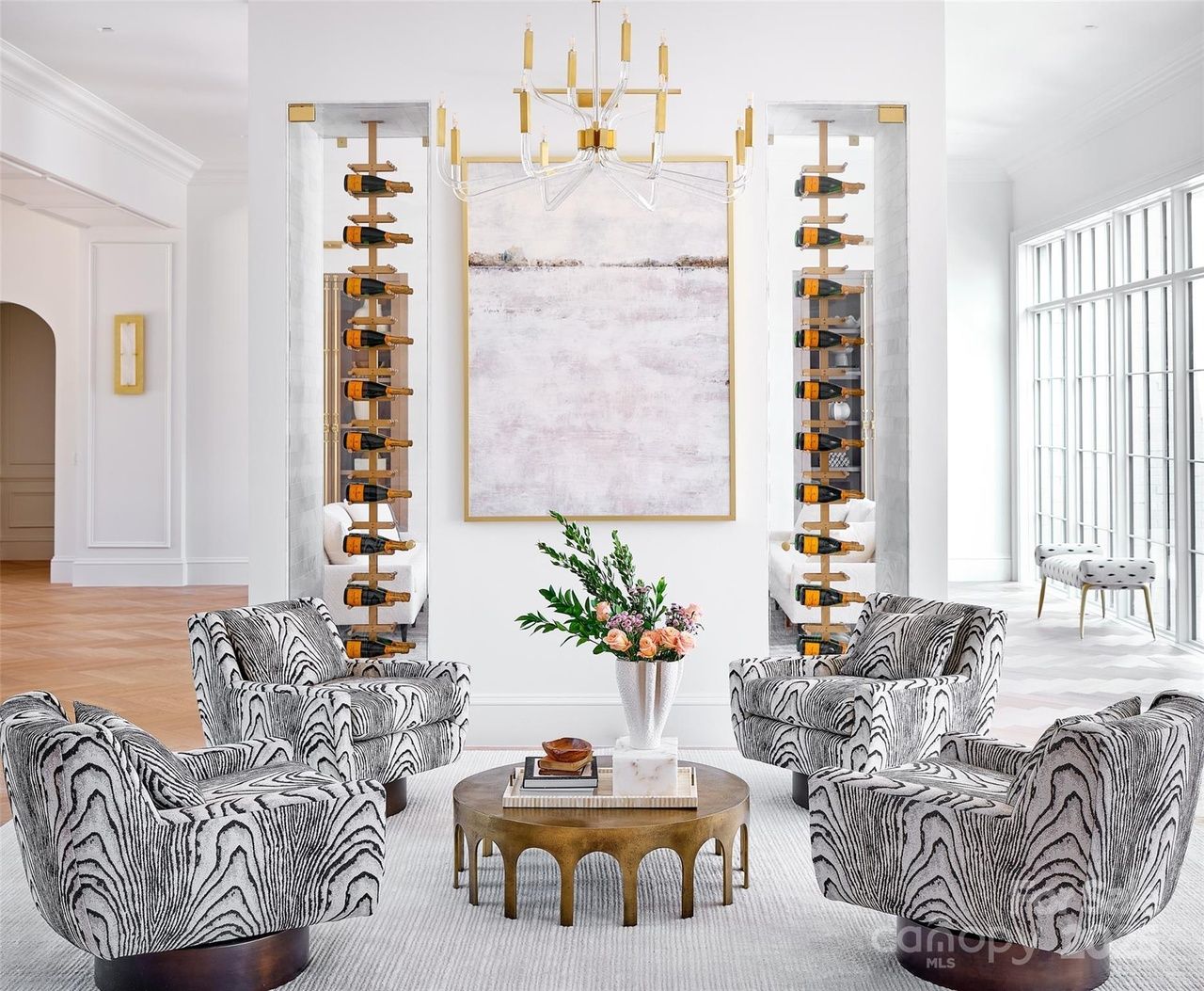 Bright living room with four patterned armchairs around a round gold coffee table holding books and flowers, white walls with two vertical wine racks and a large abstract painting.