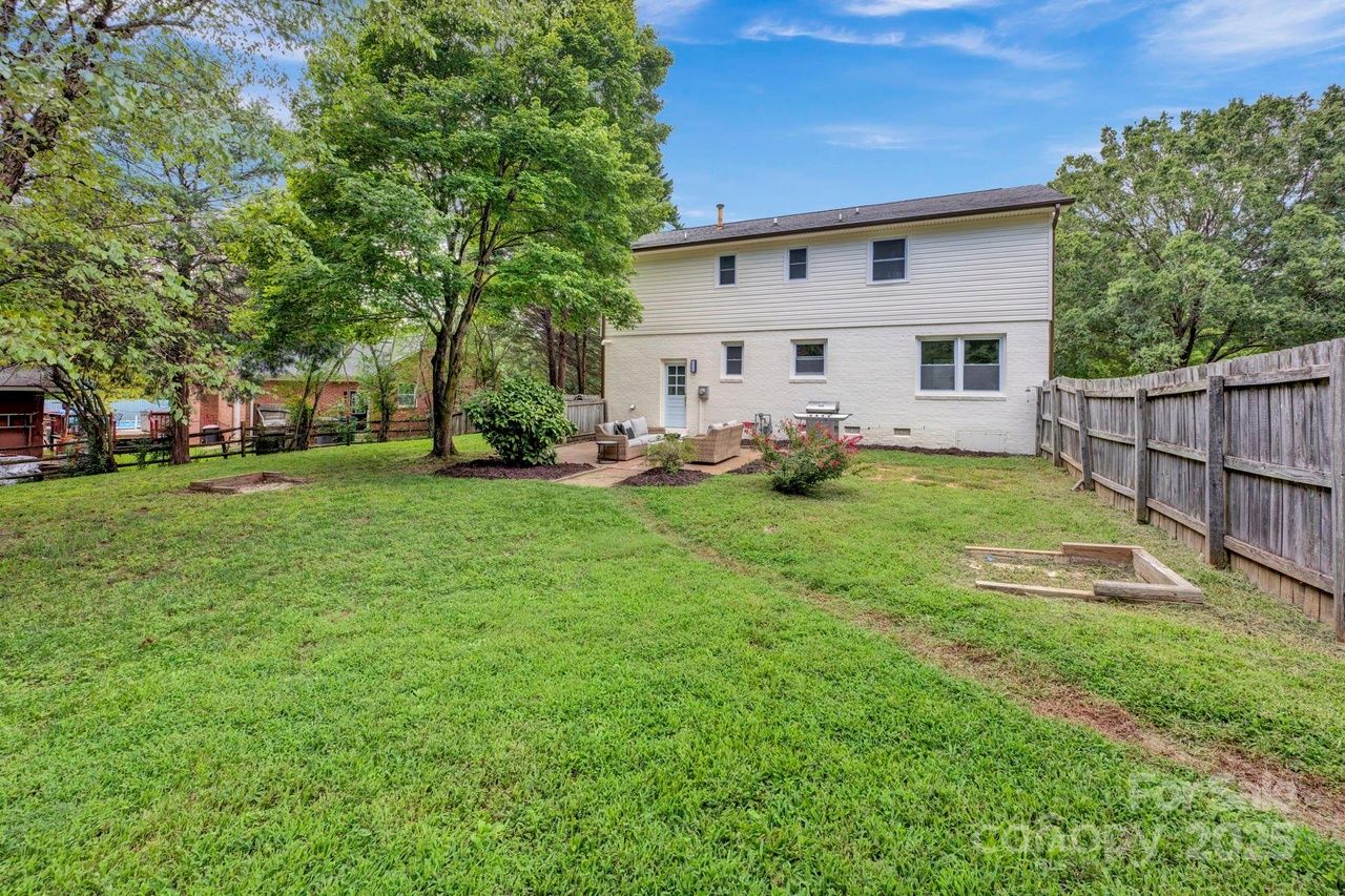 Backyard with green grass, a large tree, wooden fence, patio with outdoor sofa set, and a two-story beige house under a blue sky.