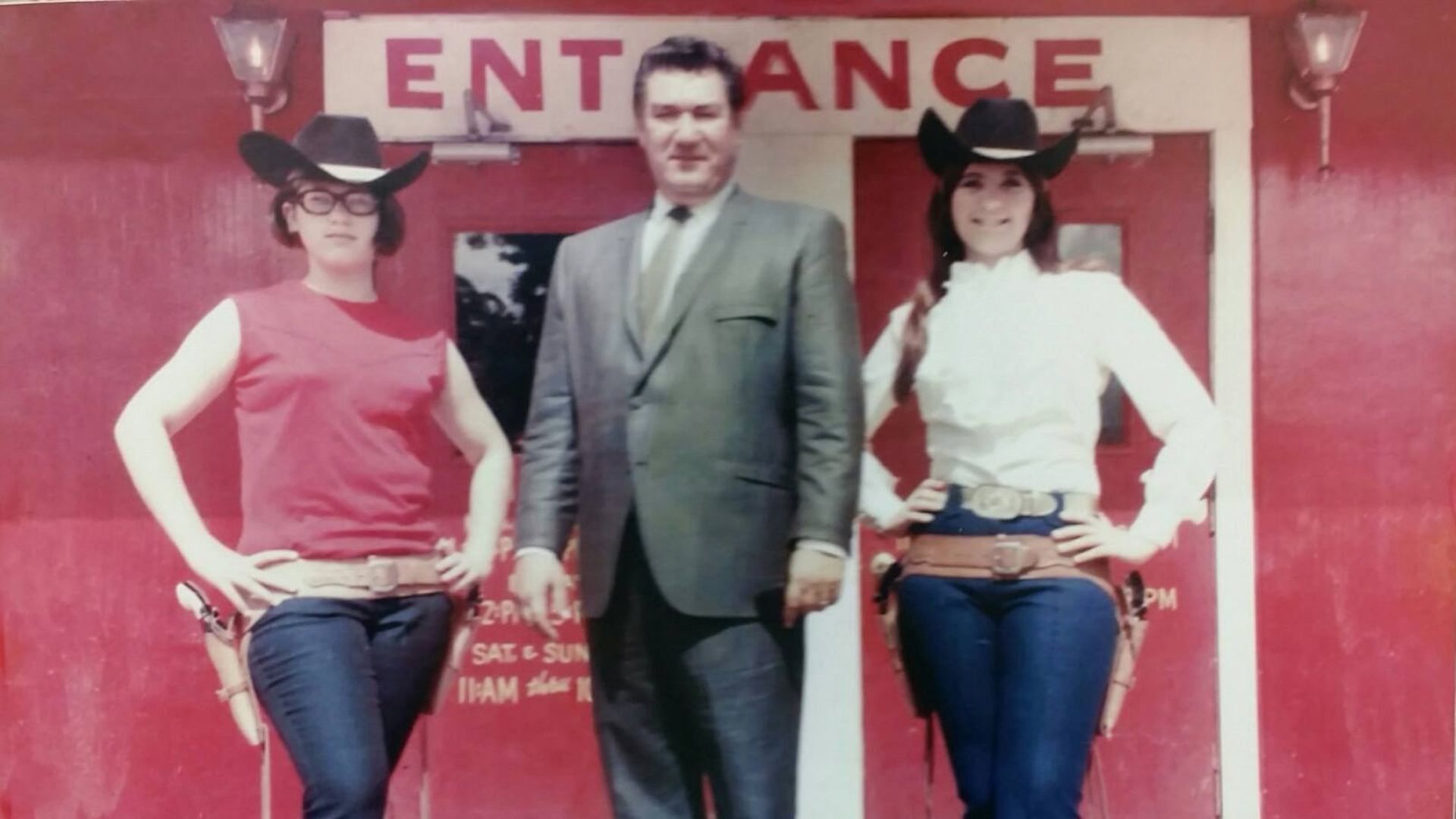 Two women, wearing western-themed outfits, to the left and right of a man in a suit. 