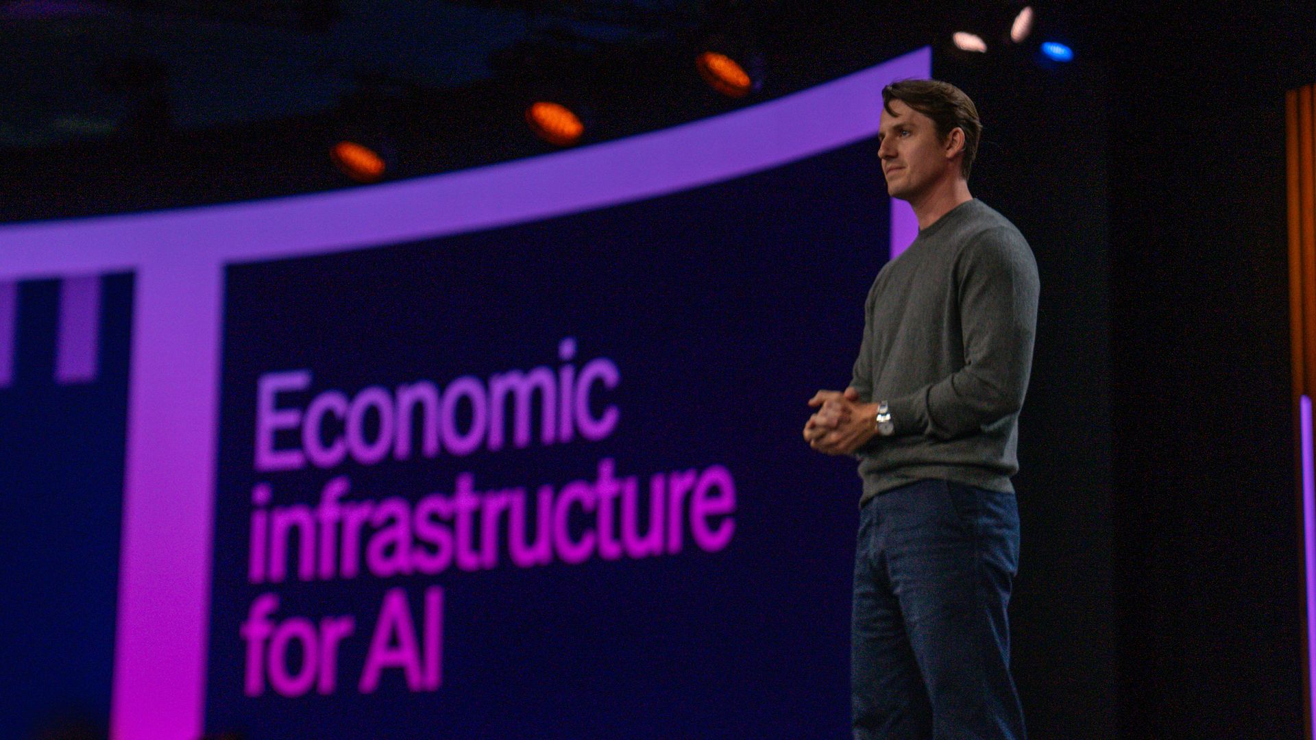 Stripe president Will Gaybrick stands on a stage beside a purple backdrop showing the pink phrase "Economic infrastructure for AI" during a presentation.