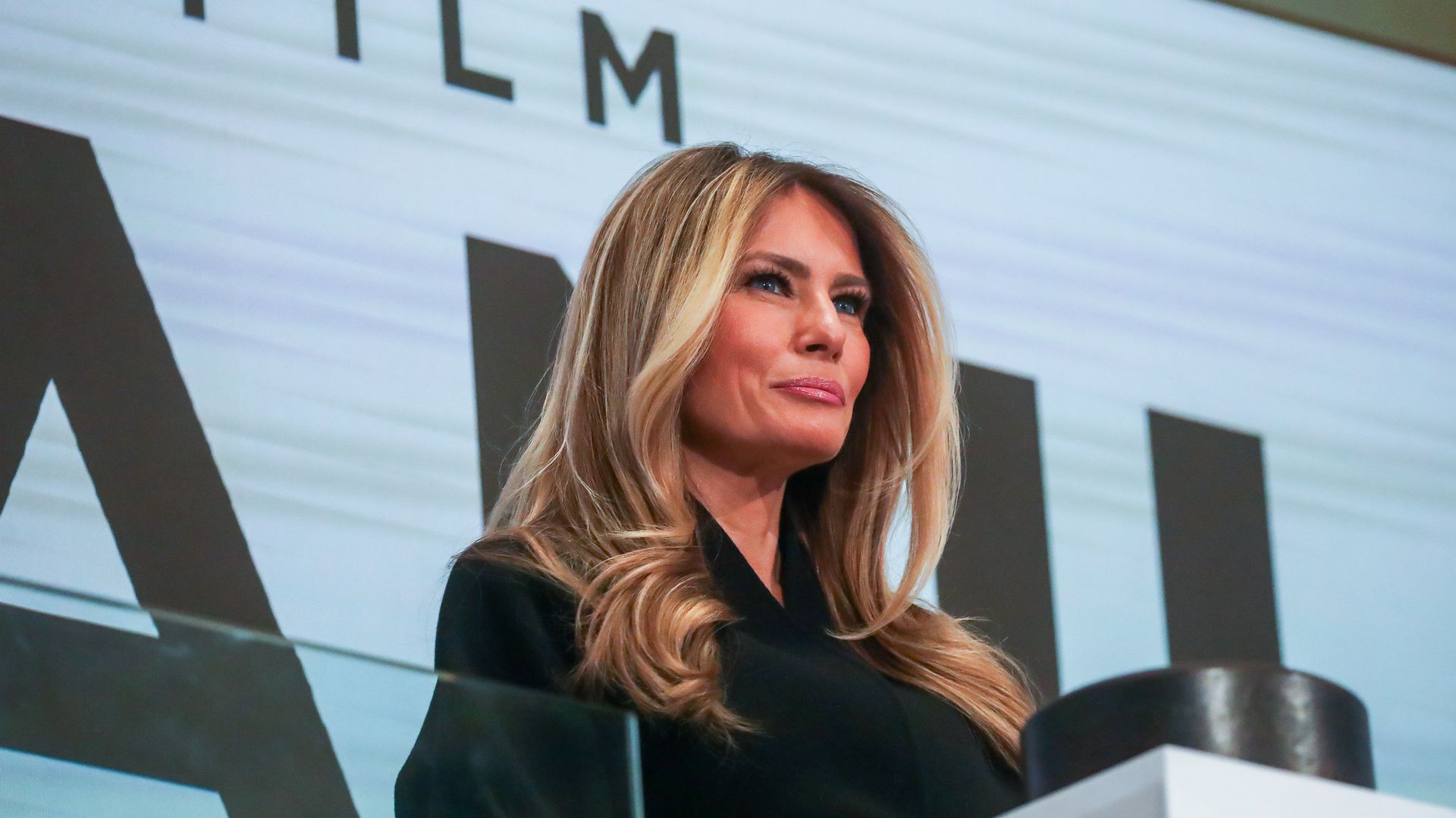 Melania Trump in black outfit with long wavy hair stands in front of a sign partly reading "FILM" and large black letters, looking thoughtfully to her right.