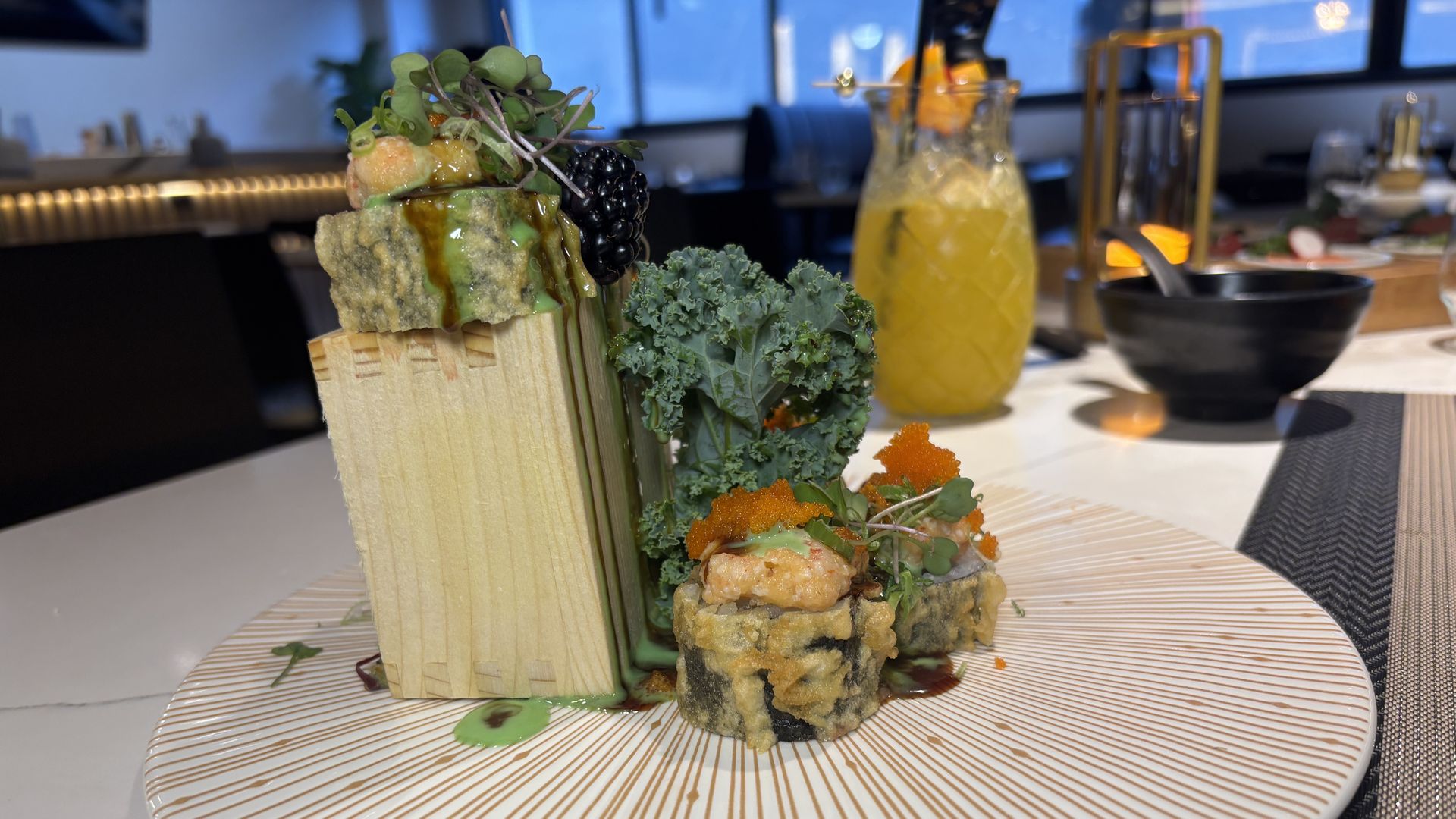 Two pieces of sushi on a white plate with orange caviar on top and greens on the side and a yellow drink in the background.
