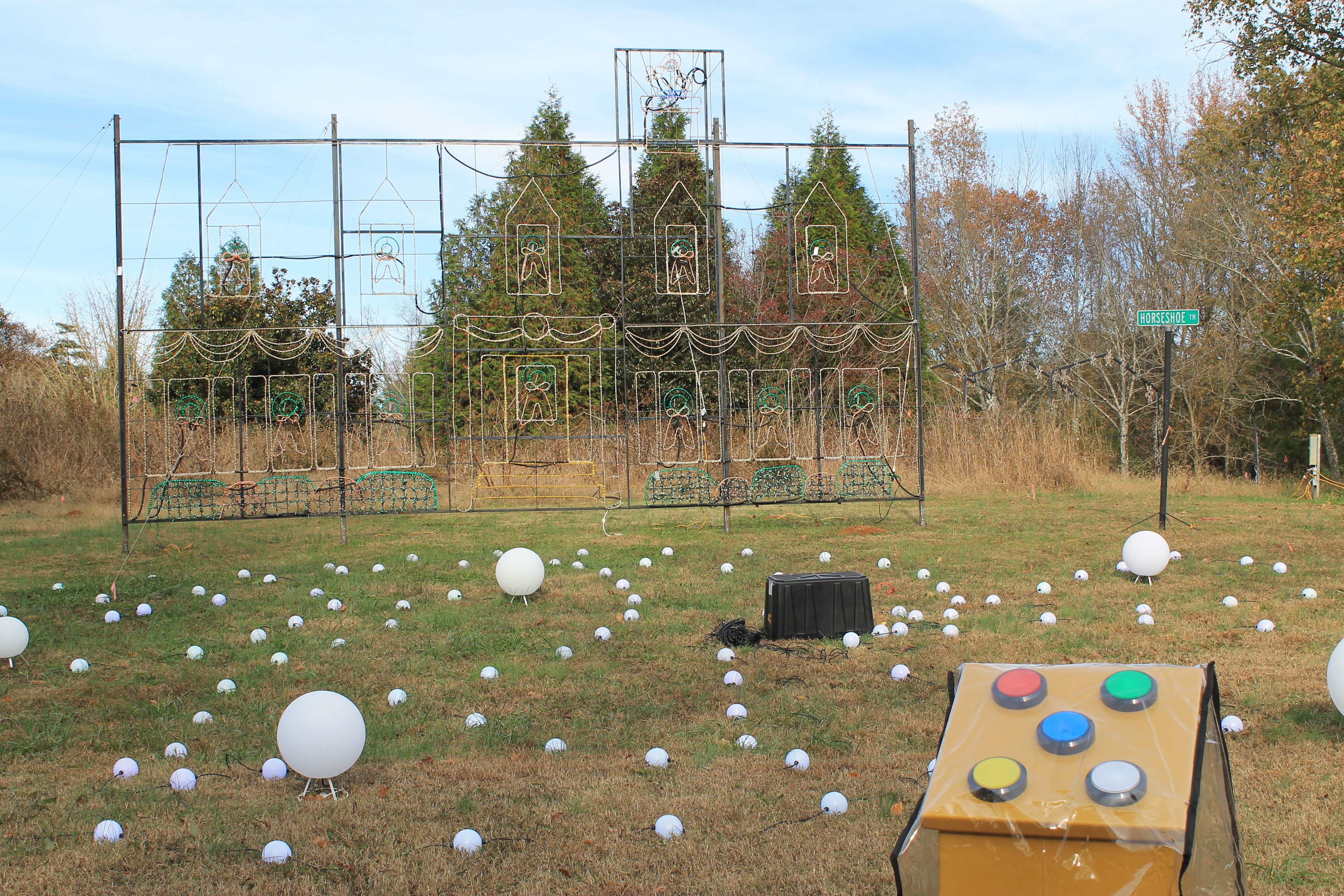 Outdoor holiday light display on grass with large colored buttons in front, white spheres scattered around, and a wire frame depicting a house with gingerbread figures in front of trees under blue sky.