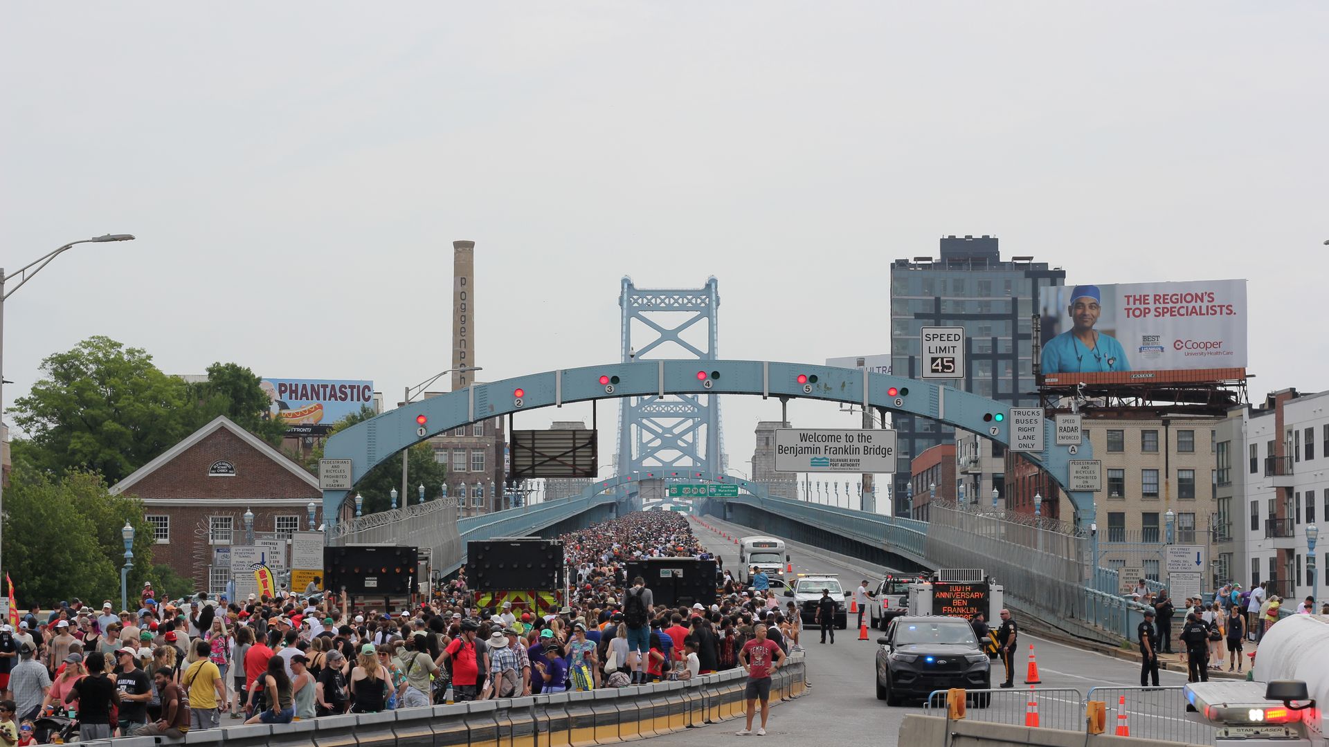 Large crowd of runners crosses a blue suspension bridge on a sunny day, with barriers, cones, and signage. City buildings and billboards line the riverfront; a police car is in the foreground.