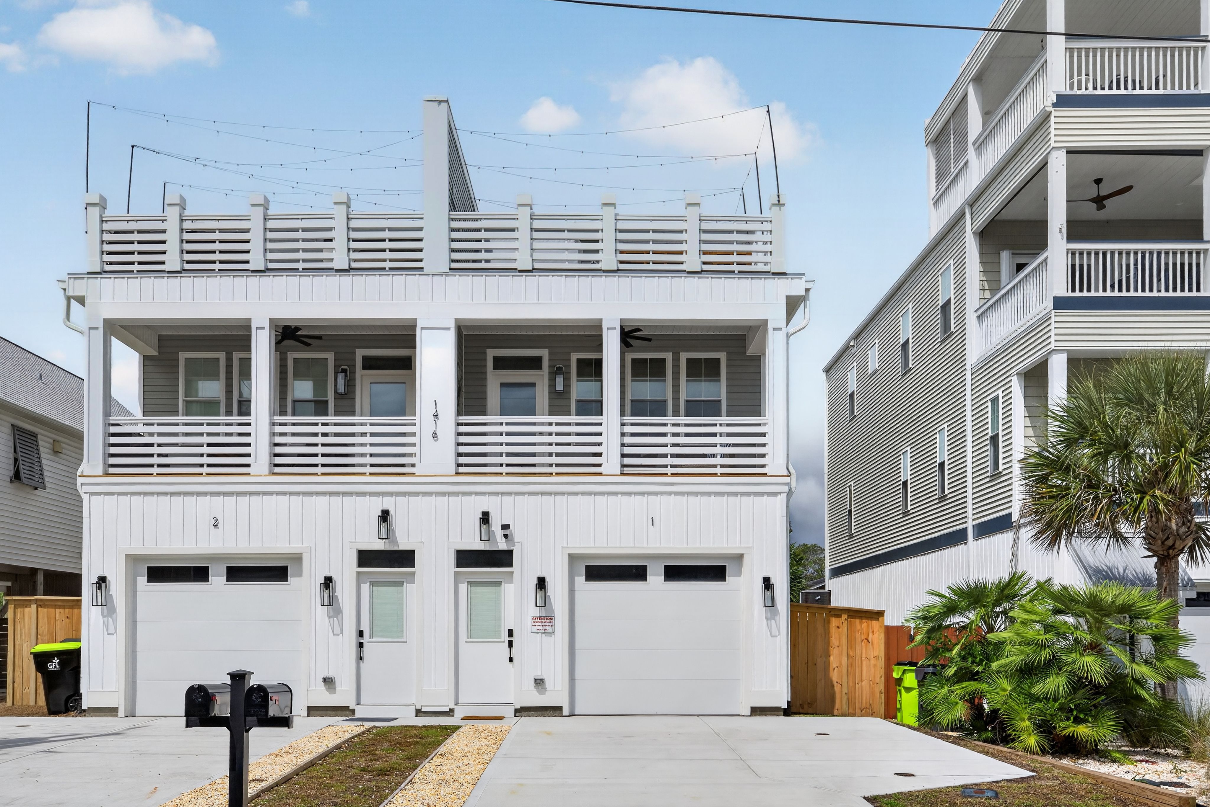 Two white coastal townhouses with multiple decks and slatted railings; string lights span the upper terrace. A palm tree stands to the right against a blue sky.