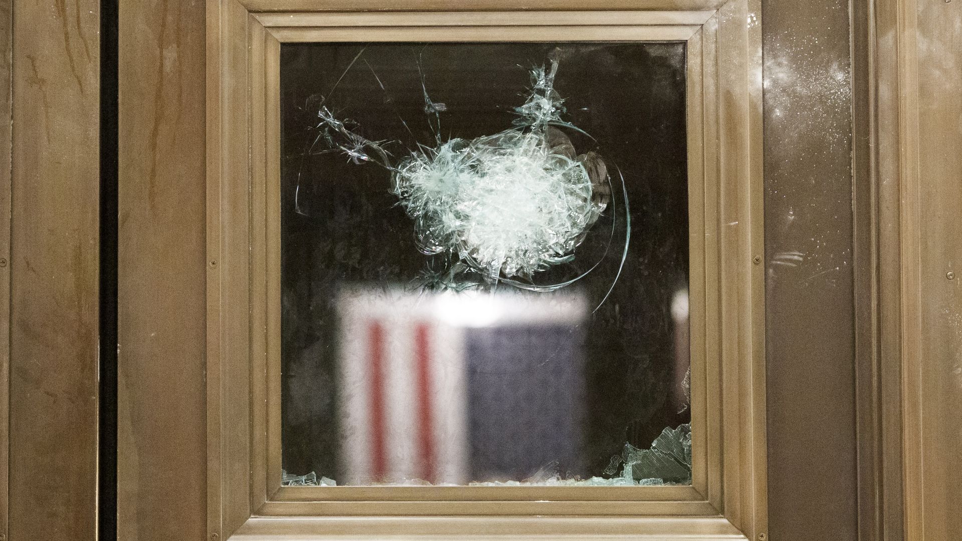 Photo of a cracked glass window looking into a room with the American flag hanging from the wall
