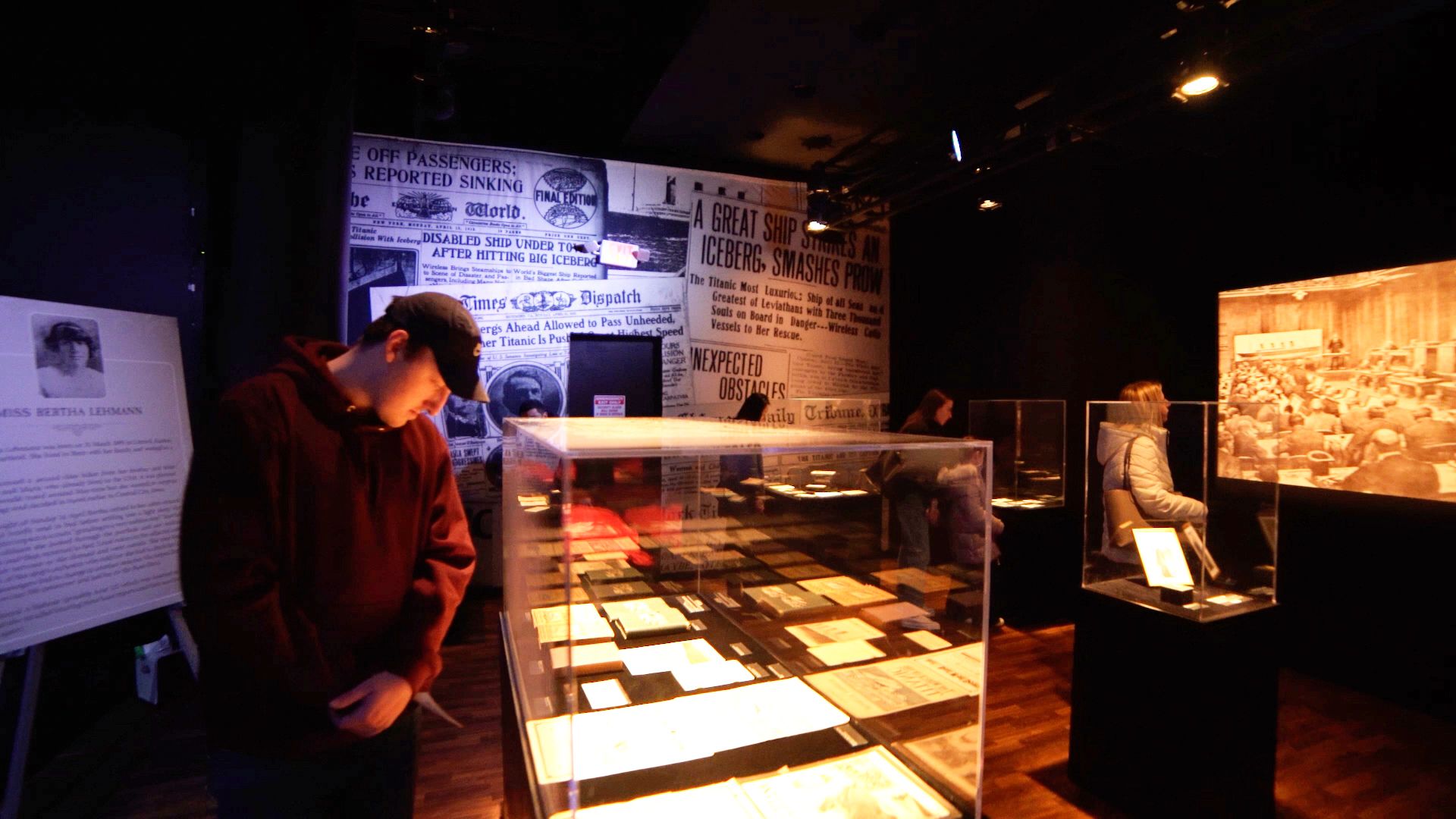 Dim museum gallery with glass display cases of manuscripts and newspapers. A young man in a maroon hoodie leans over a case; wall of old headlines; sepia-toned crowd photo on the right.