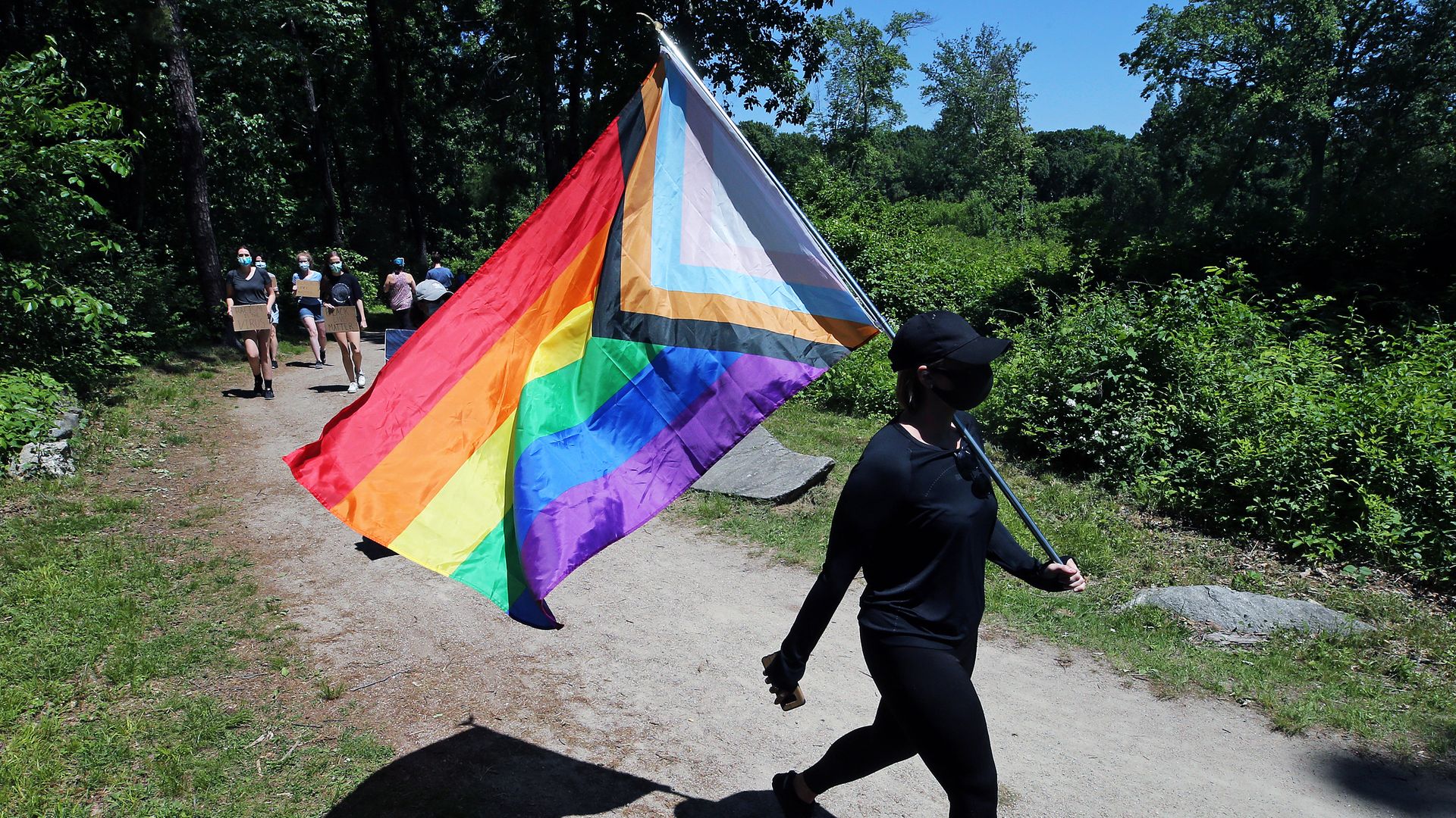 An LGBTQ flag is carried on a hiking trail surrounded by trees