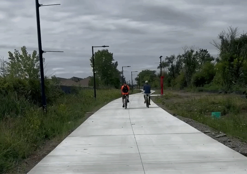 Two cyclists ride away on a straight, light-gray concrete trail under a cloudy sky. Black street lamps line both sides; green bushes and trees frame the path as it stretches into the distance.