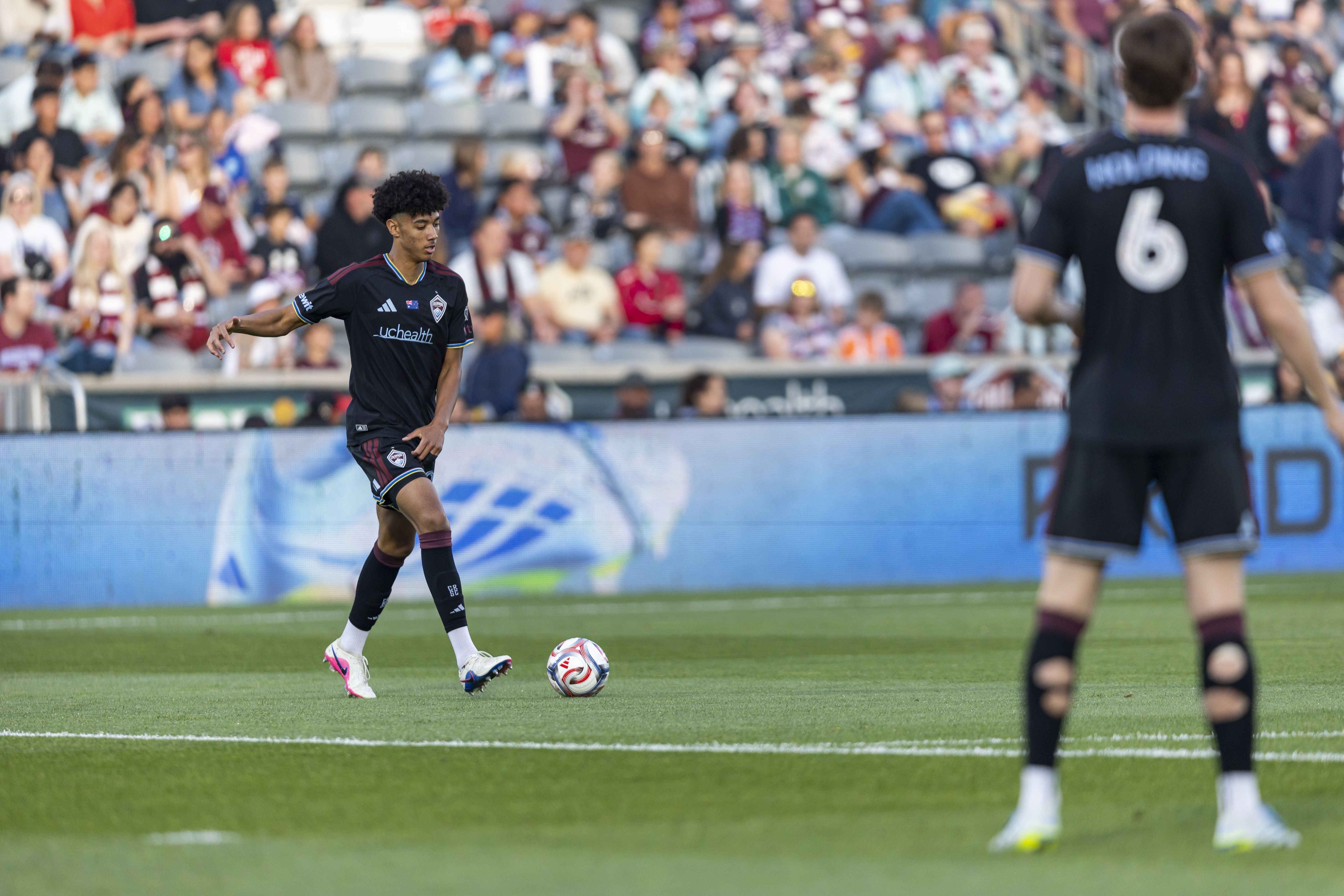 A soccer player in a dark uniform dribbles a ball on a green field, with a blurred crowd in the stands behind him and a teammate in the foreground wearing number 6.