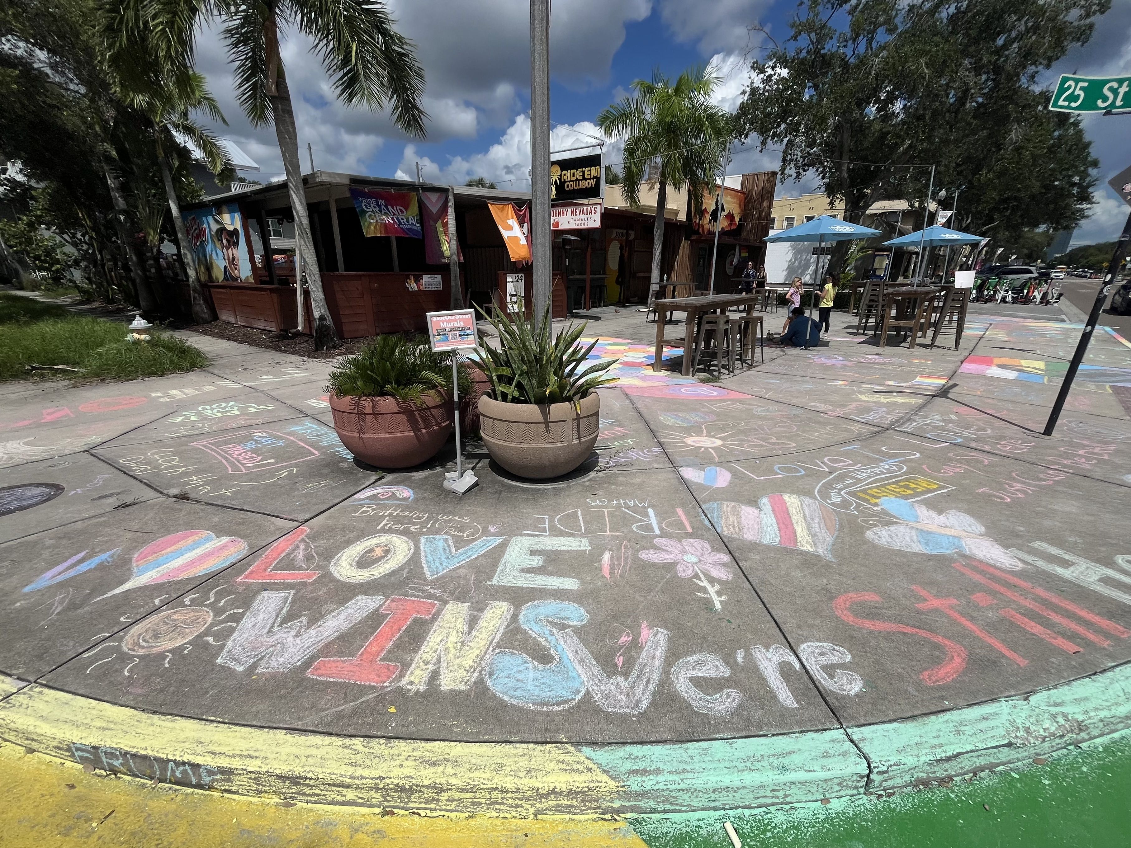 Sidewalk chalk art with colorful messages like "LOVE WINS" and rainbow hearts near palm trees and outdoor tables under blue sky at 25th Street corner.