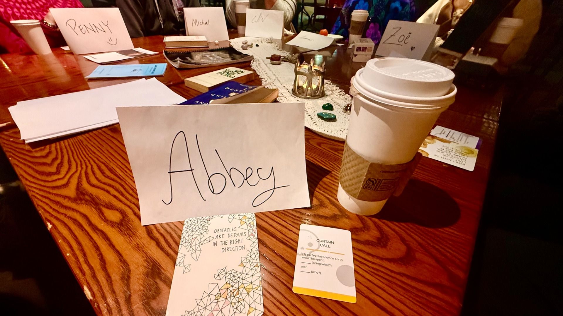 Wooden table with paper name cards for Abbey, Penny, Michael, LiV, and Zoë, a coffee cup, books, cards with inspirational text, and a lace table runner with small decorative stones and a candle holder.