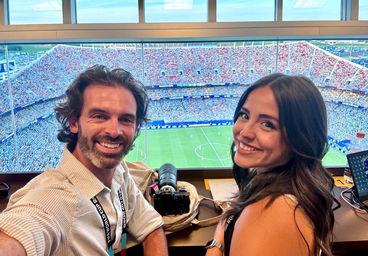 Axios reporters Travis and Abbey smiling at the camera while a camera with a long lens sits on a desk behind them and Kansas City stadium and a soccer pitch are in the background behind glass.