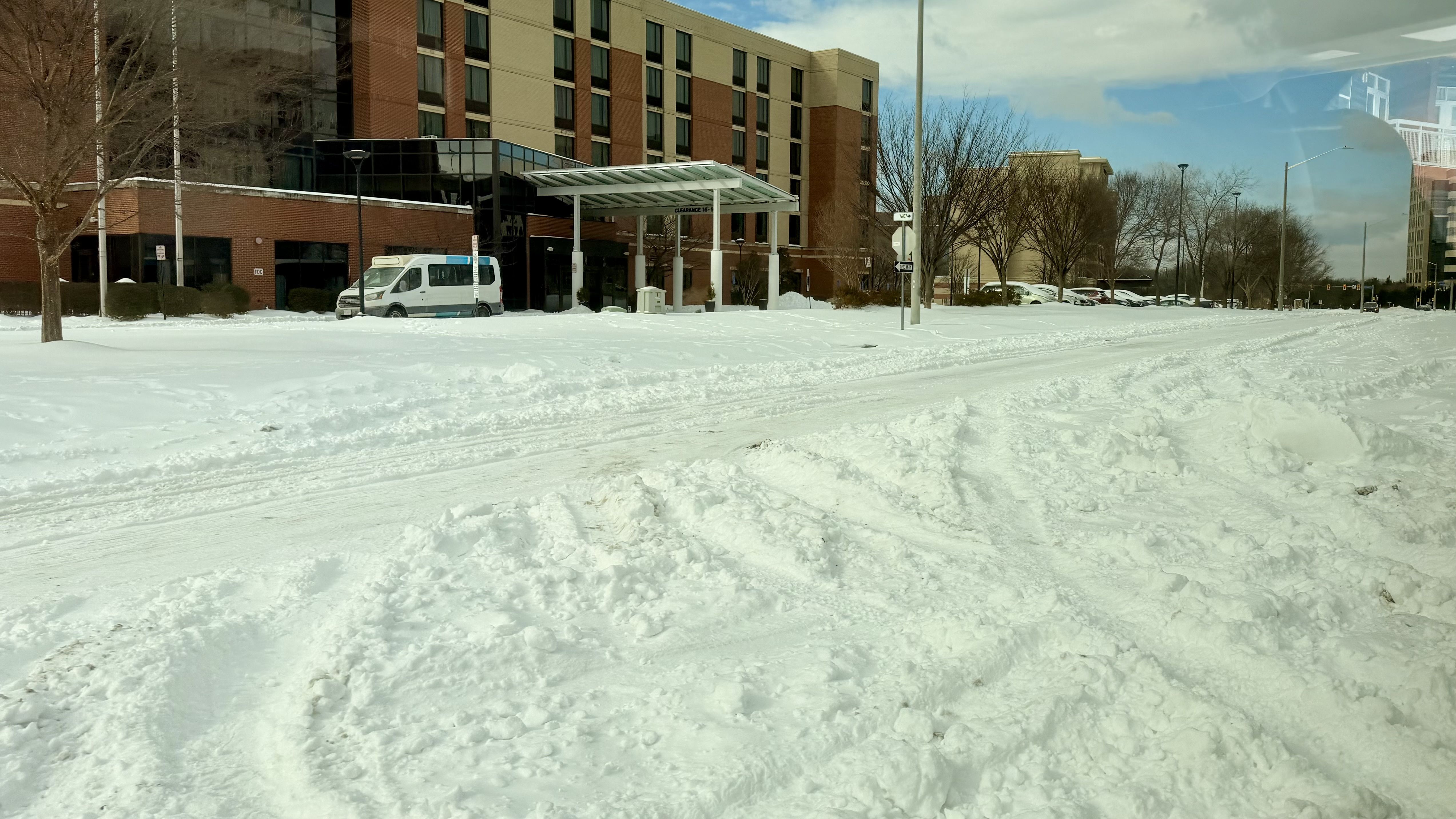 Snow-covered street and parking lot outside a brick and beige multi-story building with trees and a white van near the entrance under a glass canopy on a partly cloudy day.