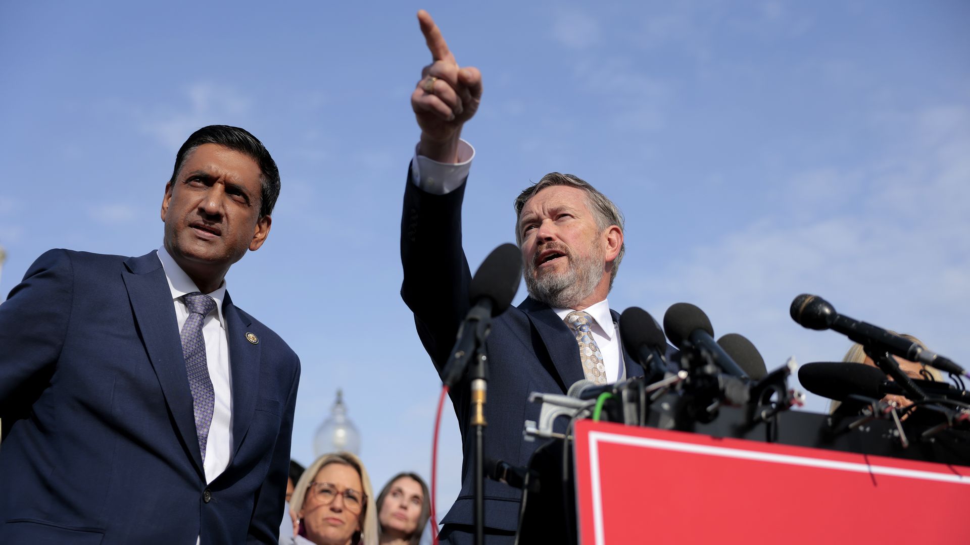Two men in suits standing at a podium with microphones, one pointing upward. Two women stand behind them. A red sign on the podium reads "EPSTEIN FILES" against a blue sky.