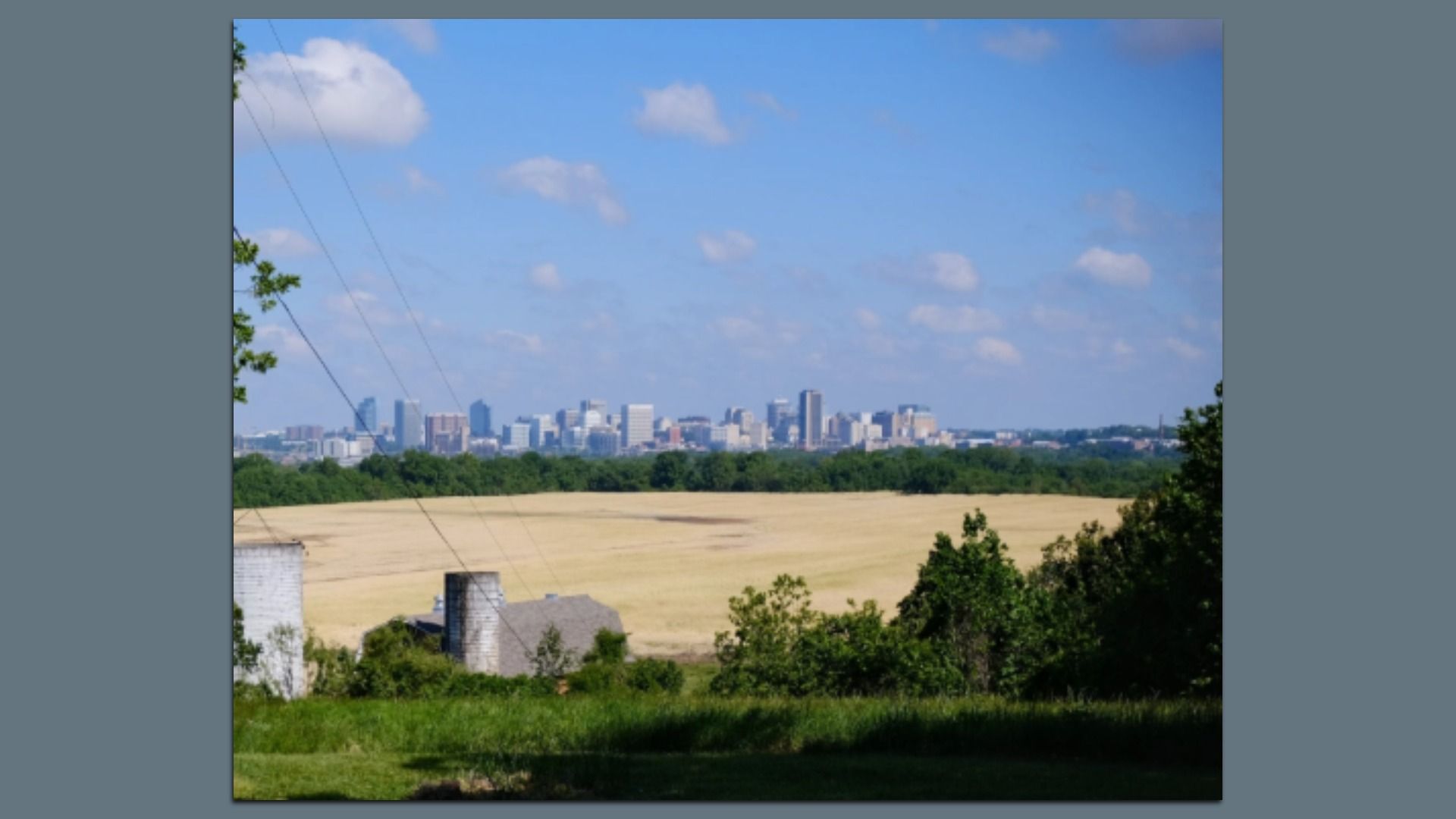 City skyline on the horizon under a blue sky with scattered white clouds; foreground shows a grassy field with trees and power lines on the left.