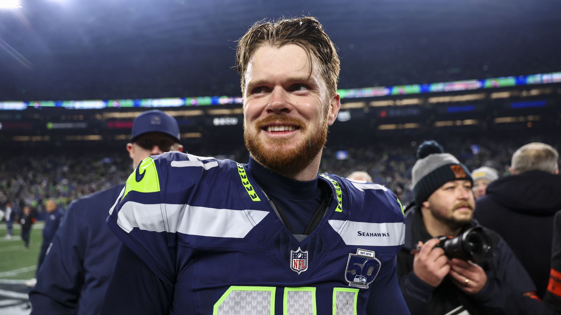 Sam Darnold, wearing a blue jersey, smiles after a football game. Behind him is a coach and a photographer. 