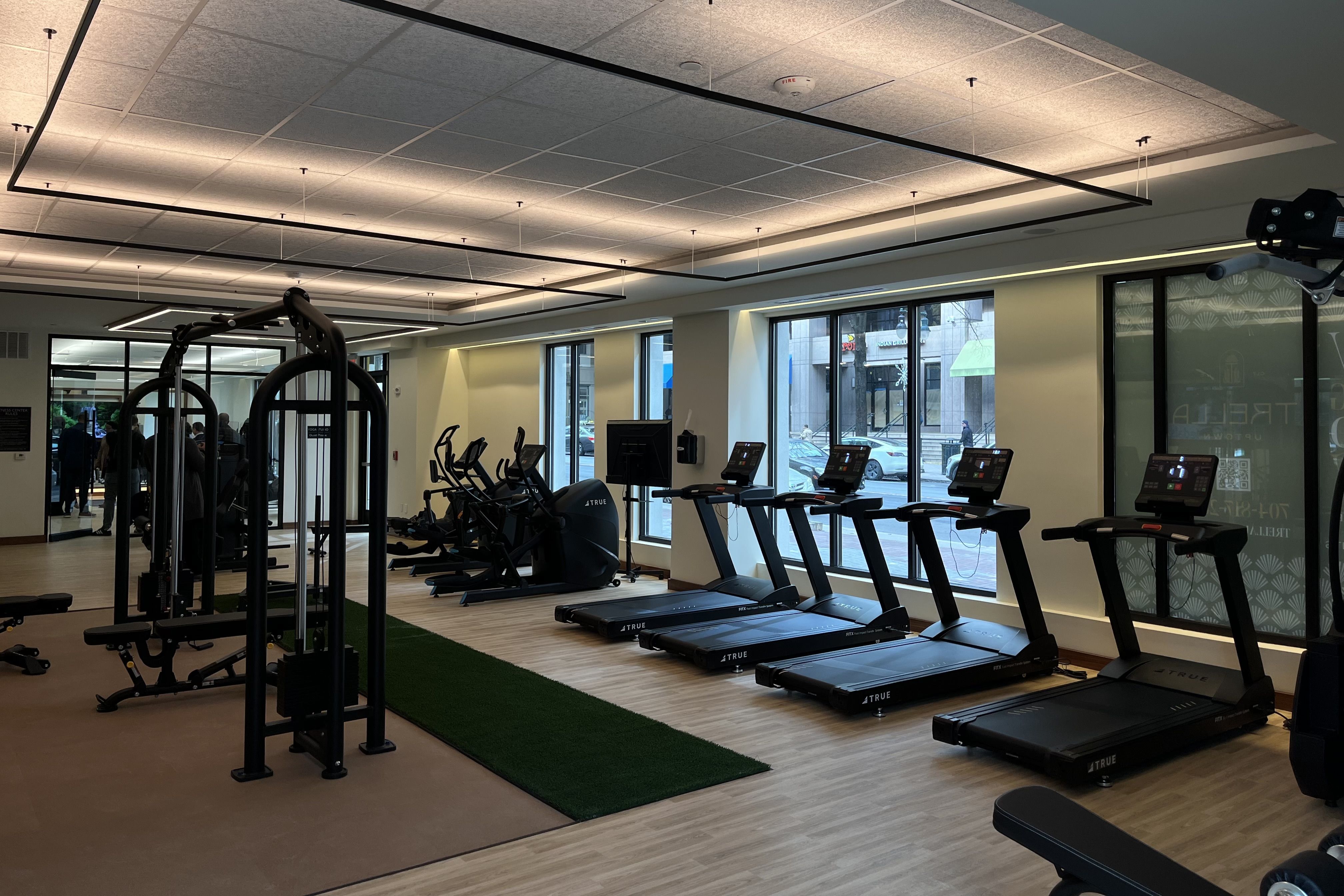 Bright gym interior with a row of black treadmills, elliptical machines, and weight machines on a wood floor next to large windows showing a city street outside.