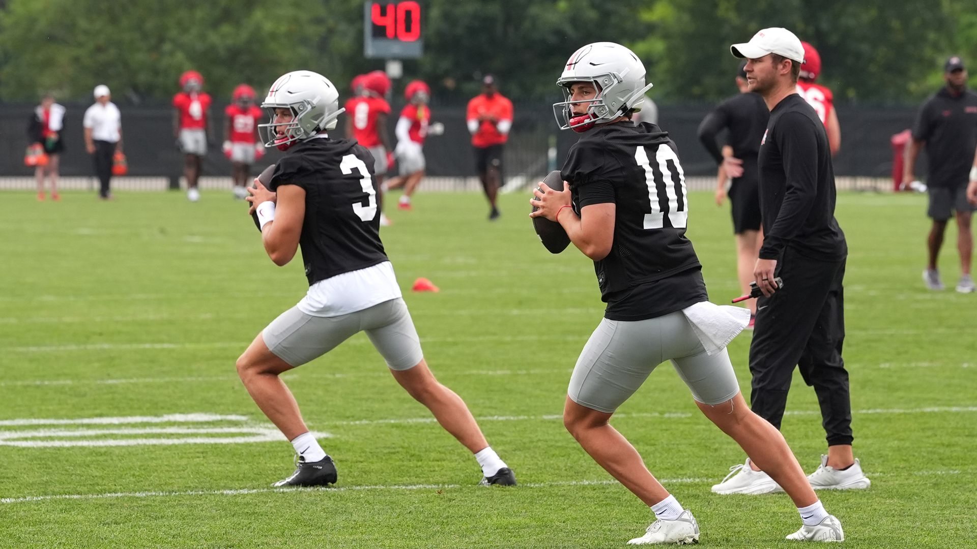 Two football players in black jerseys numbered 3 and 10 with silver helmets practice on green field, holding footballs, with a coach in black clothing and white cap nearby.