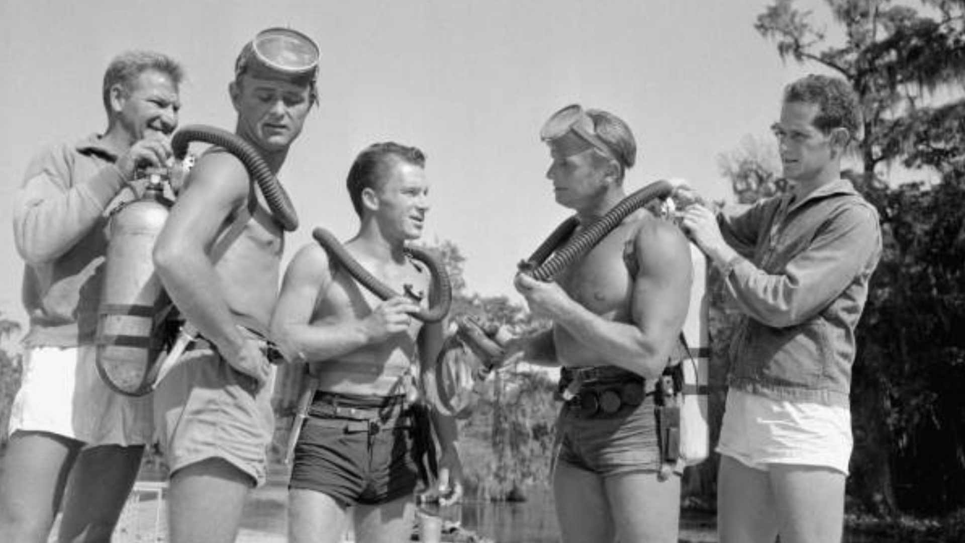 A black-and-white photo showing three shirtless men wearing diving gear and swim trunks and two men on each side of them wearing jackets helping secure the dive tanks.