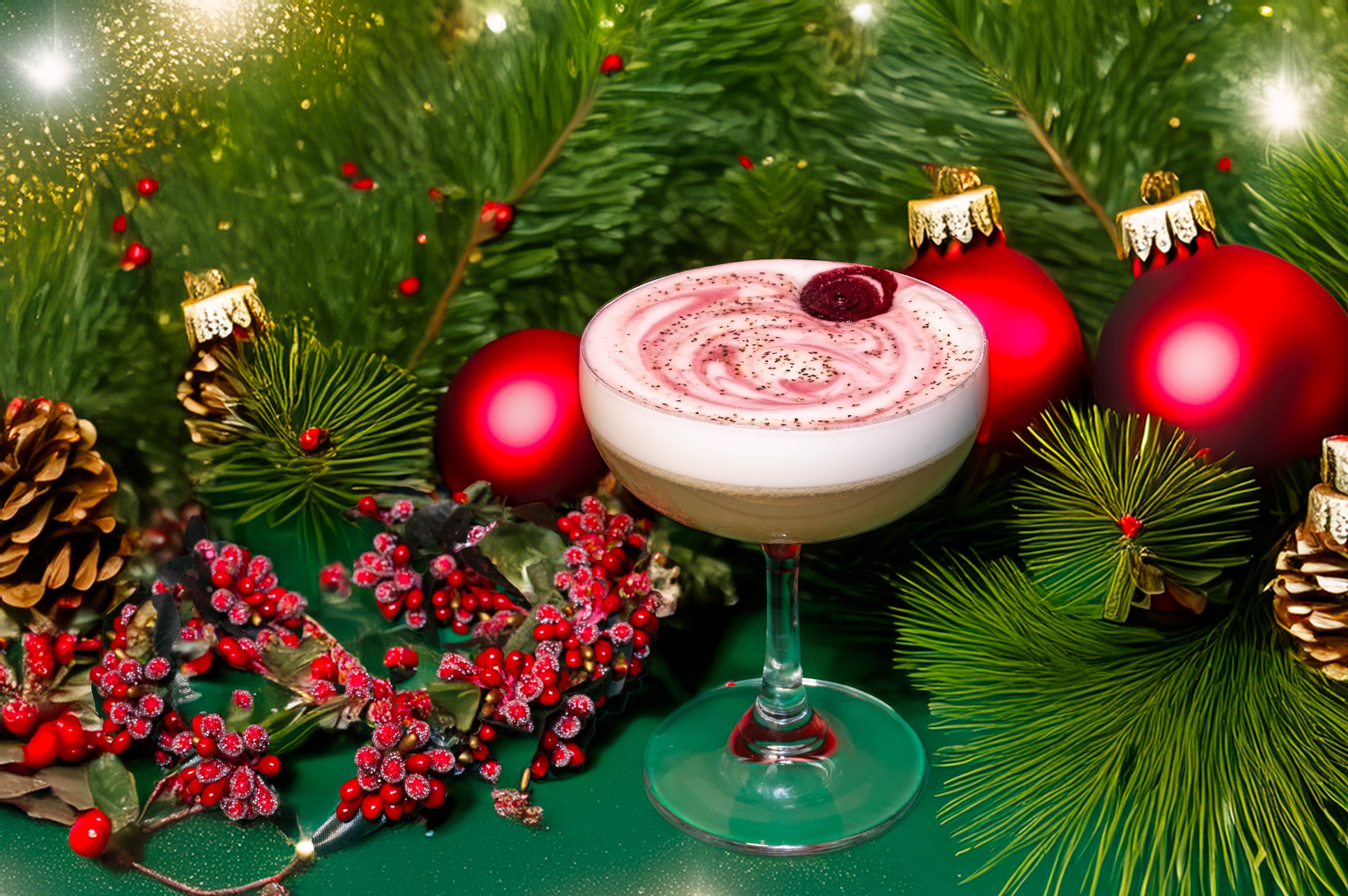 Festive cocktail with pink foam served in a glass surrounded by green pine branches, red Christmas ornaments, pine cones, and frosted red berries on a green surface.