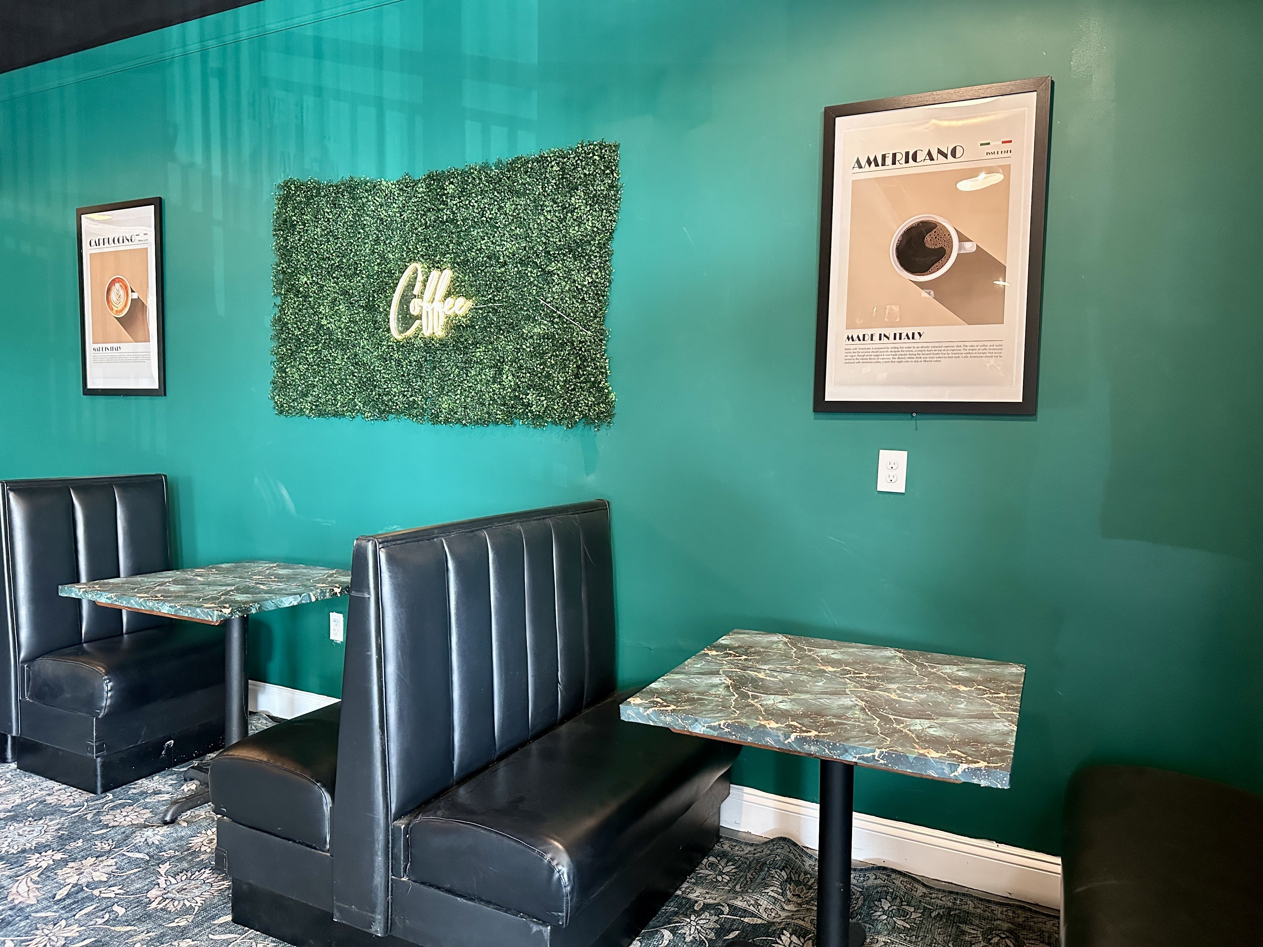Songbirds. Teal cafe with a green faux hedge wall and a glowing 'Coffee' sign. Framed posters read 'Americano' and 'Cappuccino'; black leather booths and marble tables complete the space.